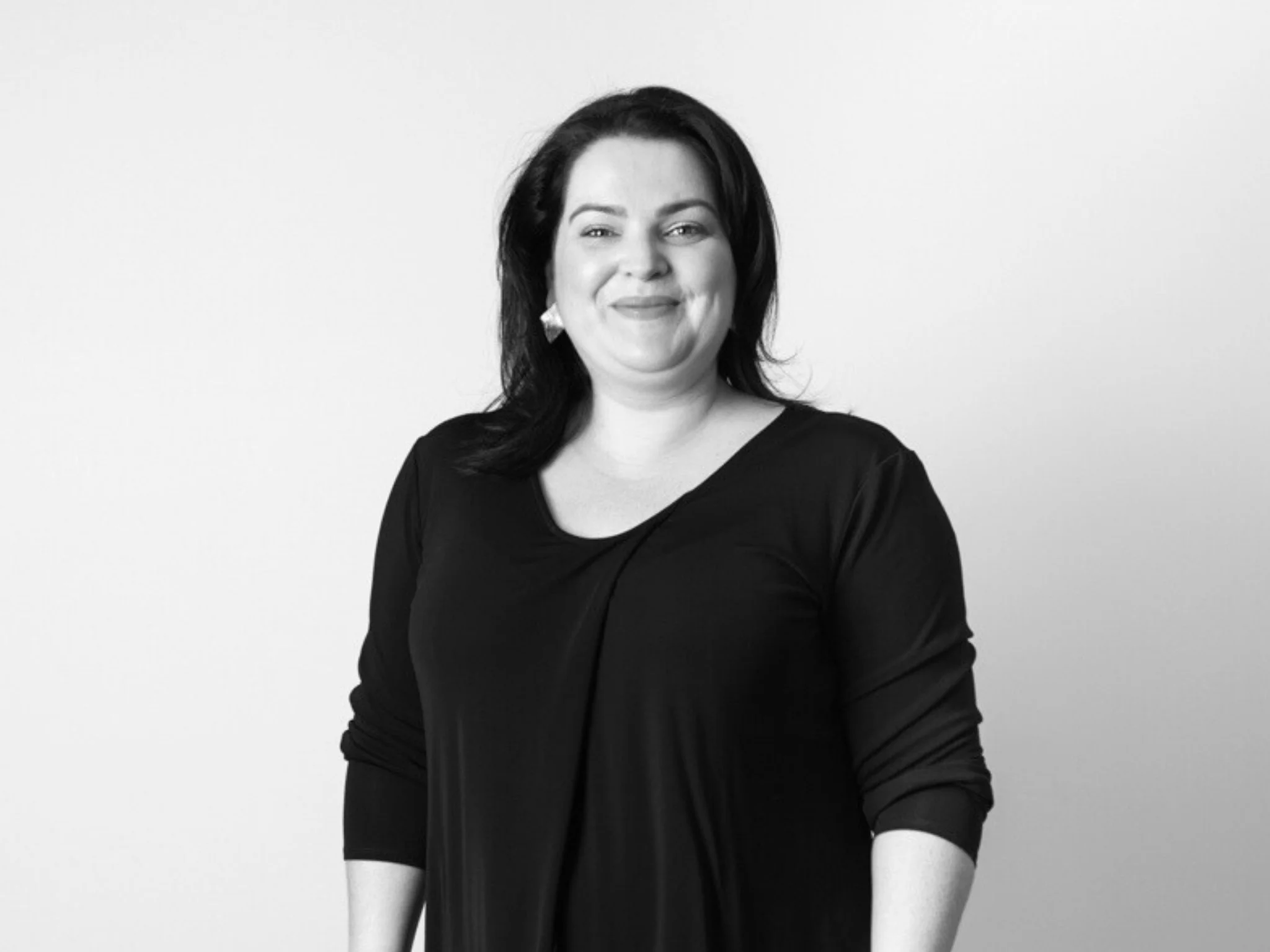 Black and white portrait of a smiling woman with dark hair, wearing a black top, standing against a plain light-colored background.