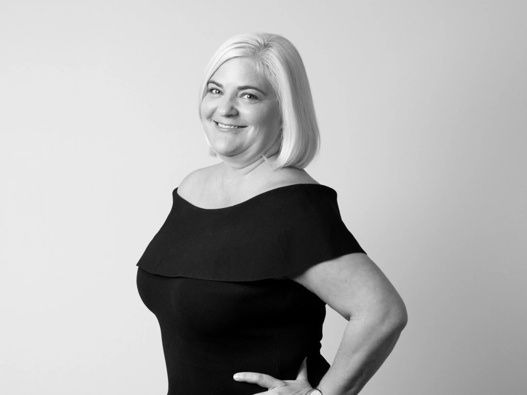 Black and white portrait of a smiling middle-aged woman with shoulder-length blonde hair, wearing an off-shoulder black dress, standing against a plain background.