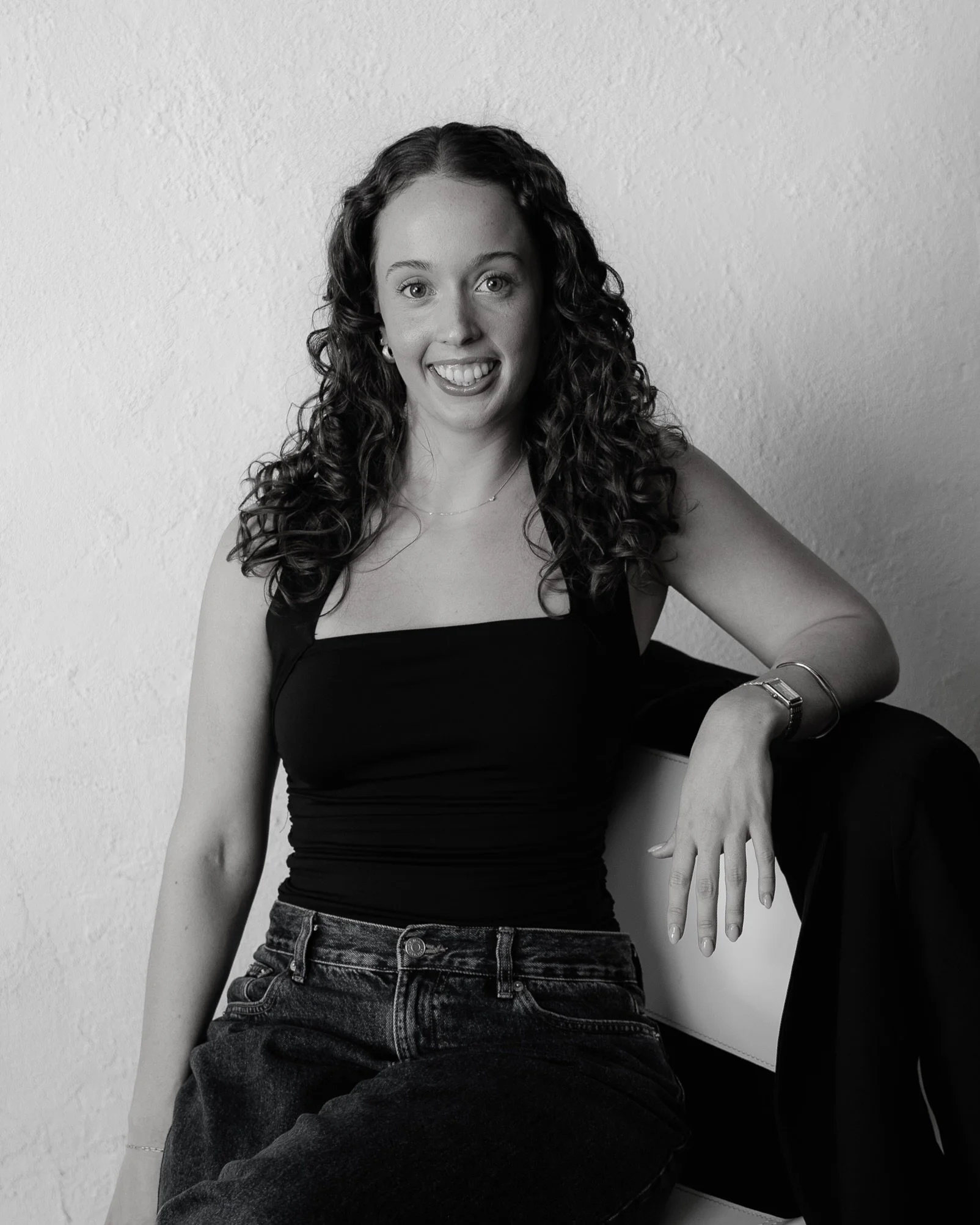 A young woman with long hair tied to one side, wearing a sleeveless black top, holding a wine glass, smiling, standing in front of a plain background.