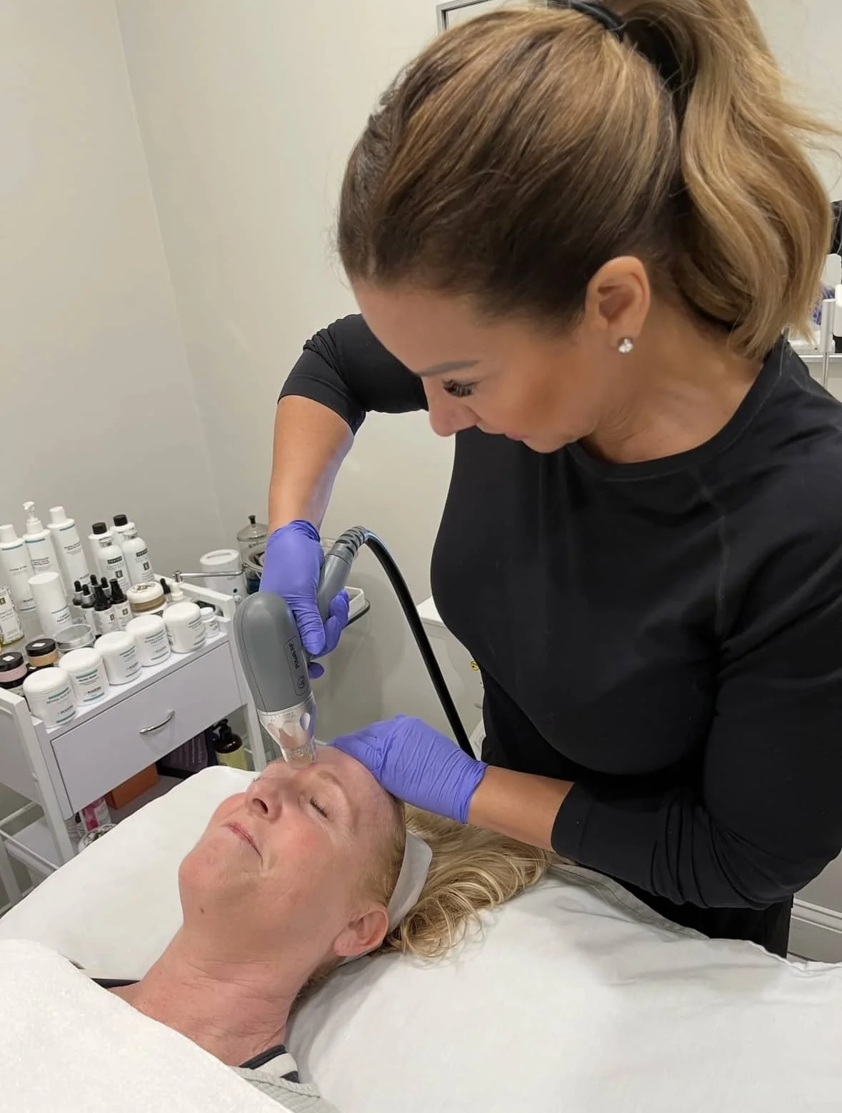 A woman in black clothing performs a facial treatment with a handheld device on a woman lying on a treatment bed, in a clinical skincare setting.