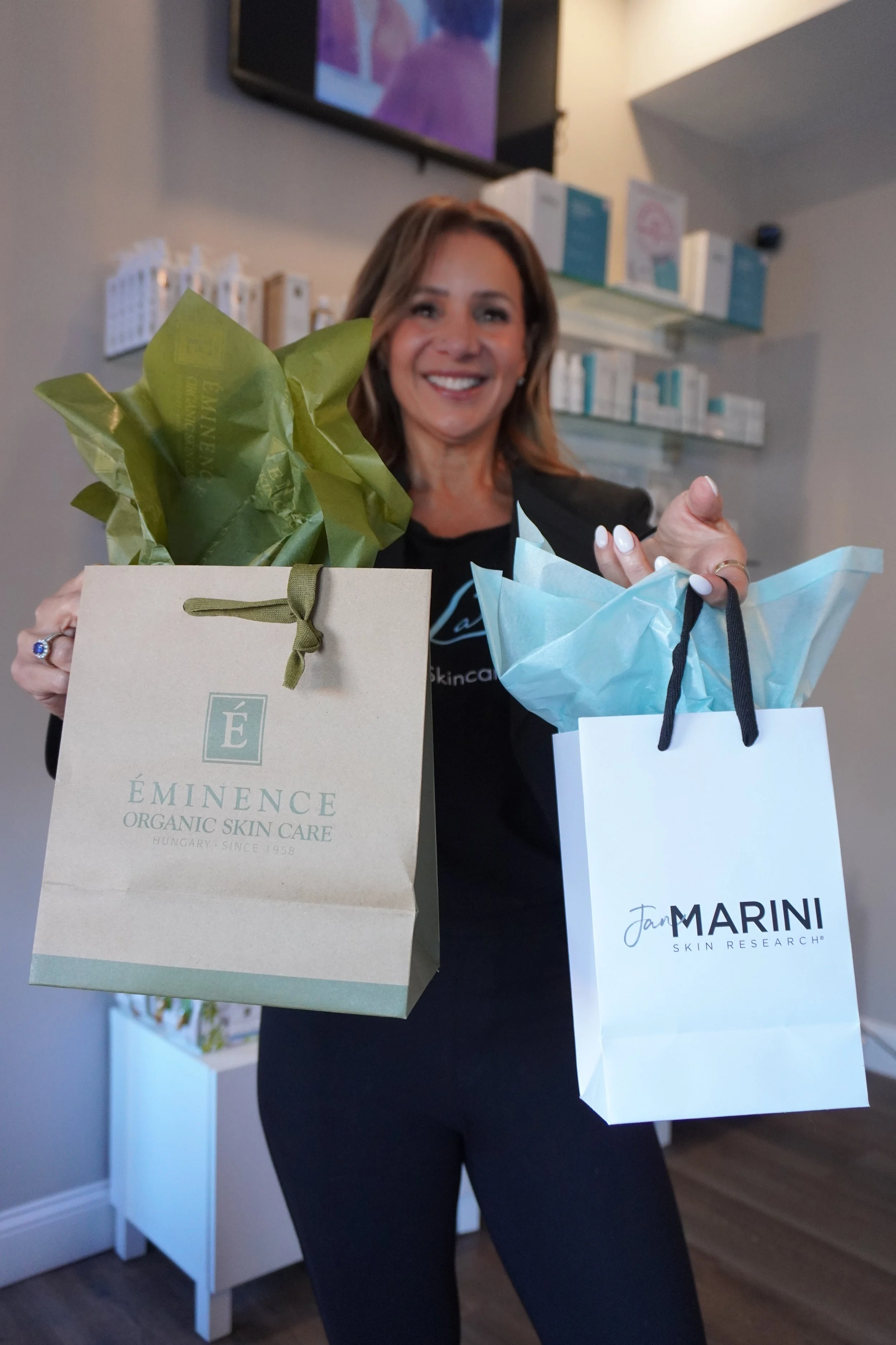 A smiling woman holds two shopping bags, one from Éminence Organic Skin Care and the other from Marini Skin Research, inside a skincare store.