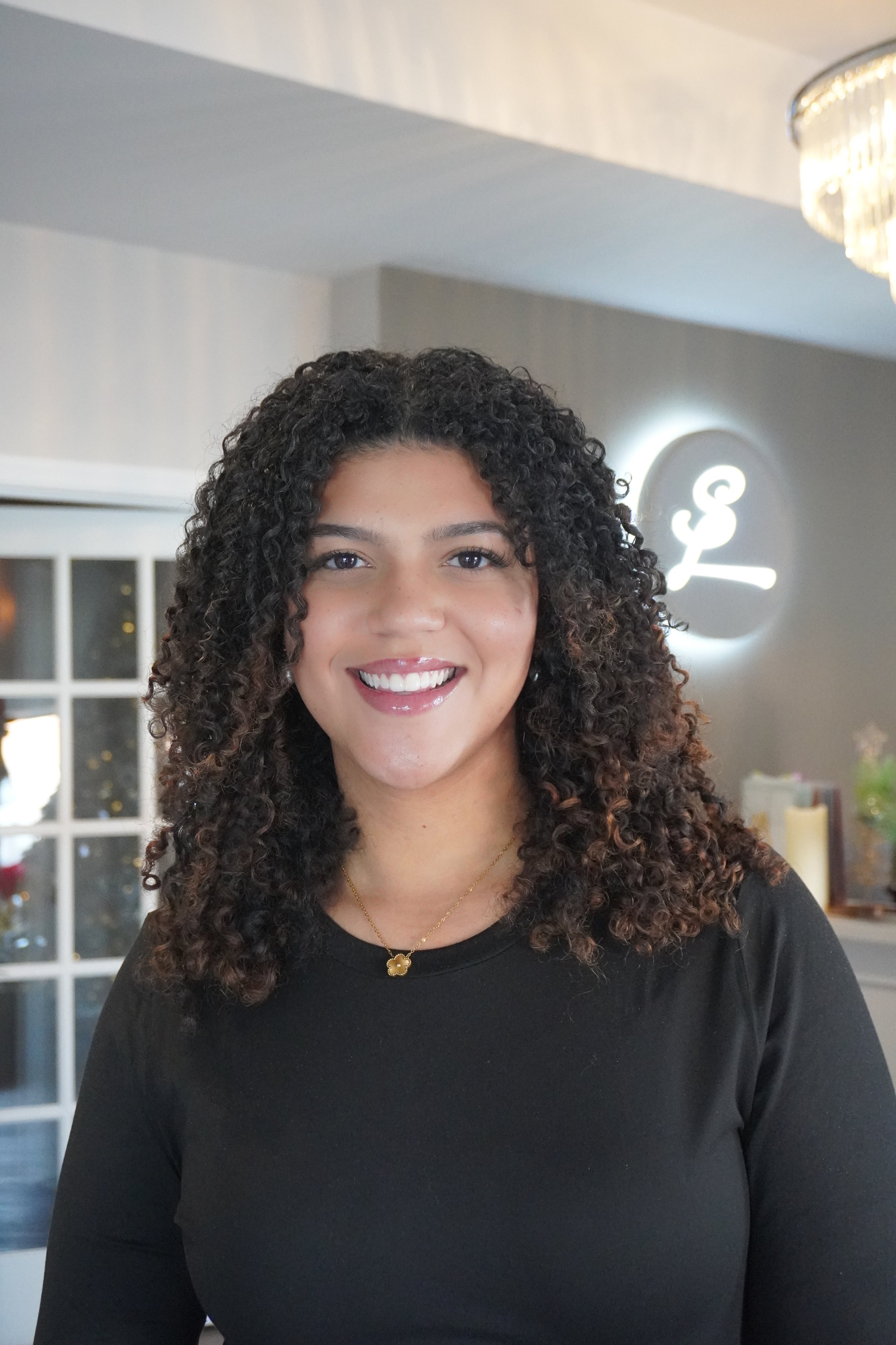Rebecca with curly dark hair and a black top smiling indoors, with a lit sign in the background.