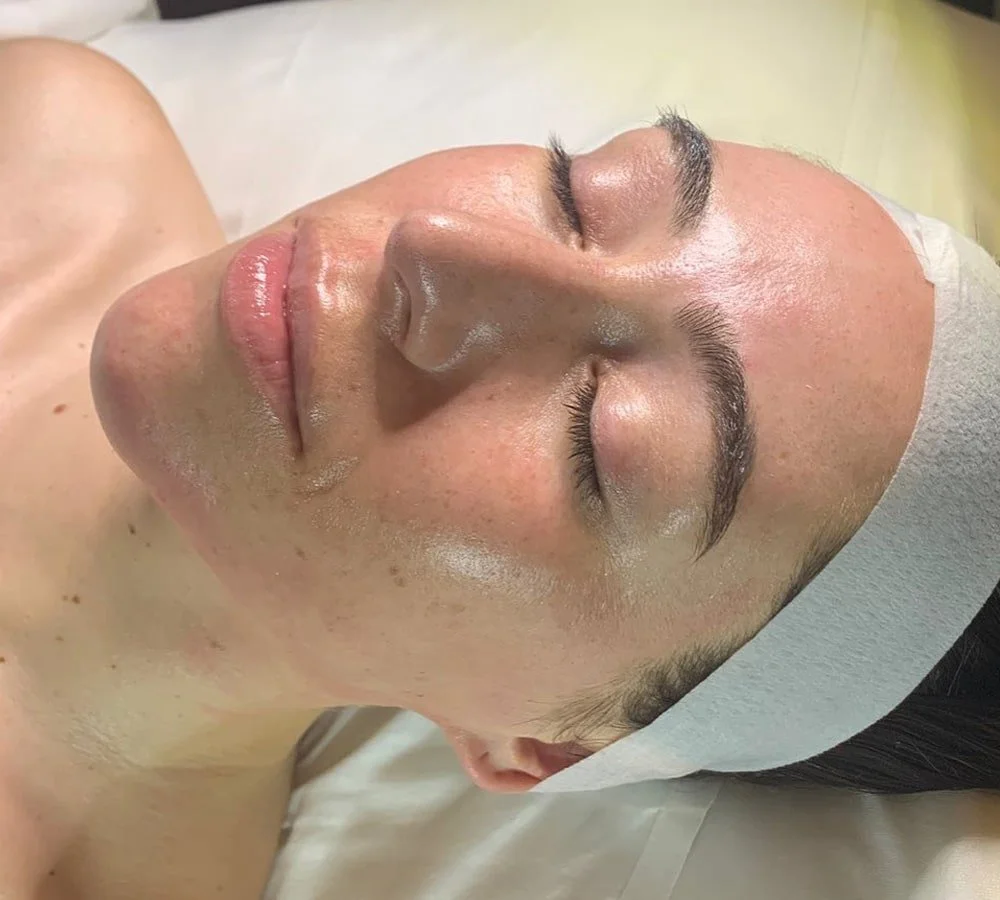 A woman lying down with her eyes closed, getting a facial treatment. She has a headband around her hair to keep it away from her face.
