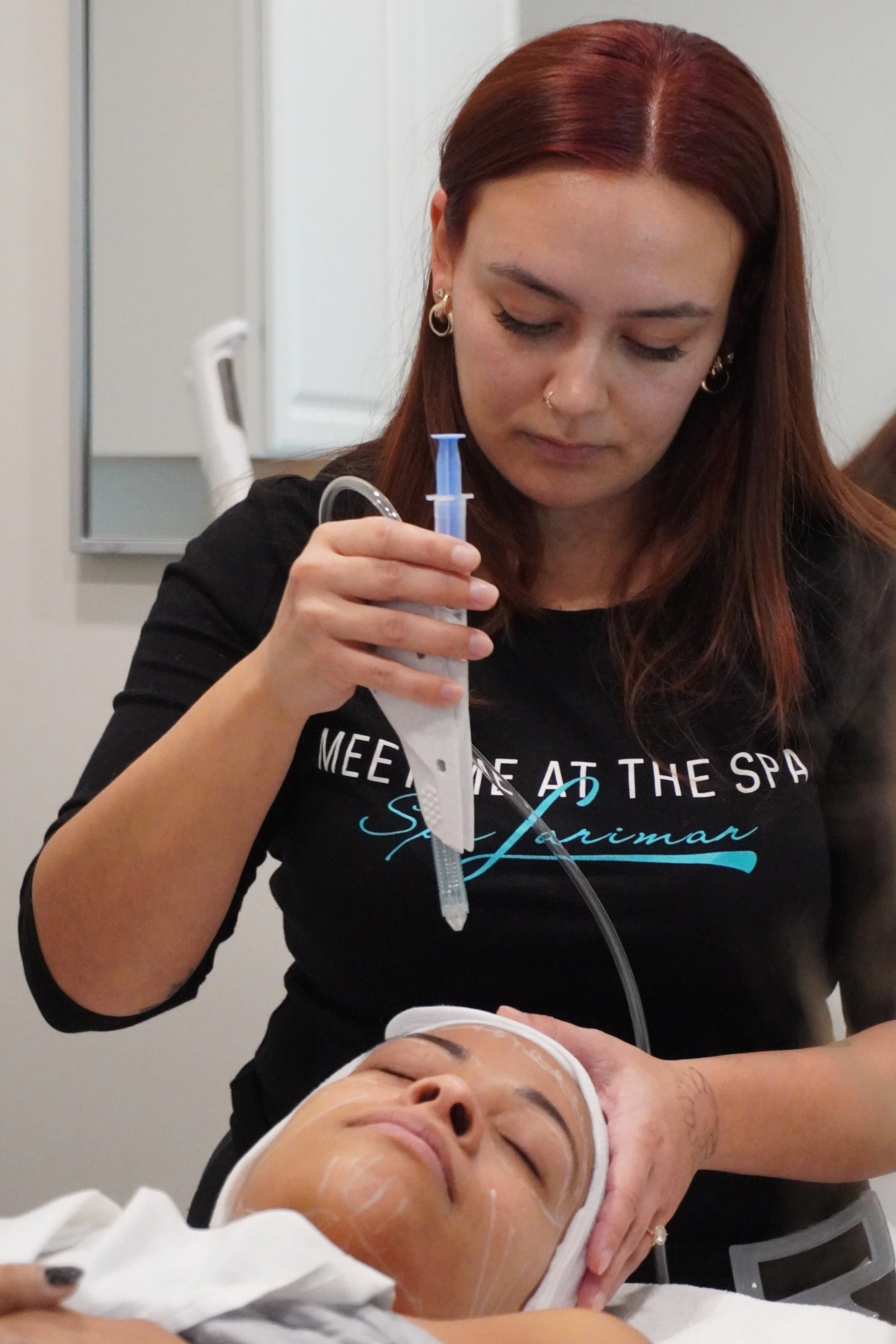 A woman with red hair performing a facial treatment on a woman lying down with her eyes closed. The aesthetician is holding a syringe-like device with a tube, likely applying a facial treatment or extracting.
