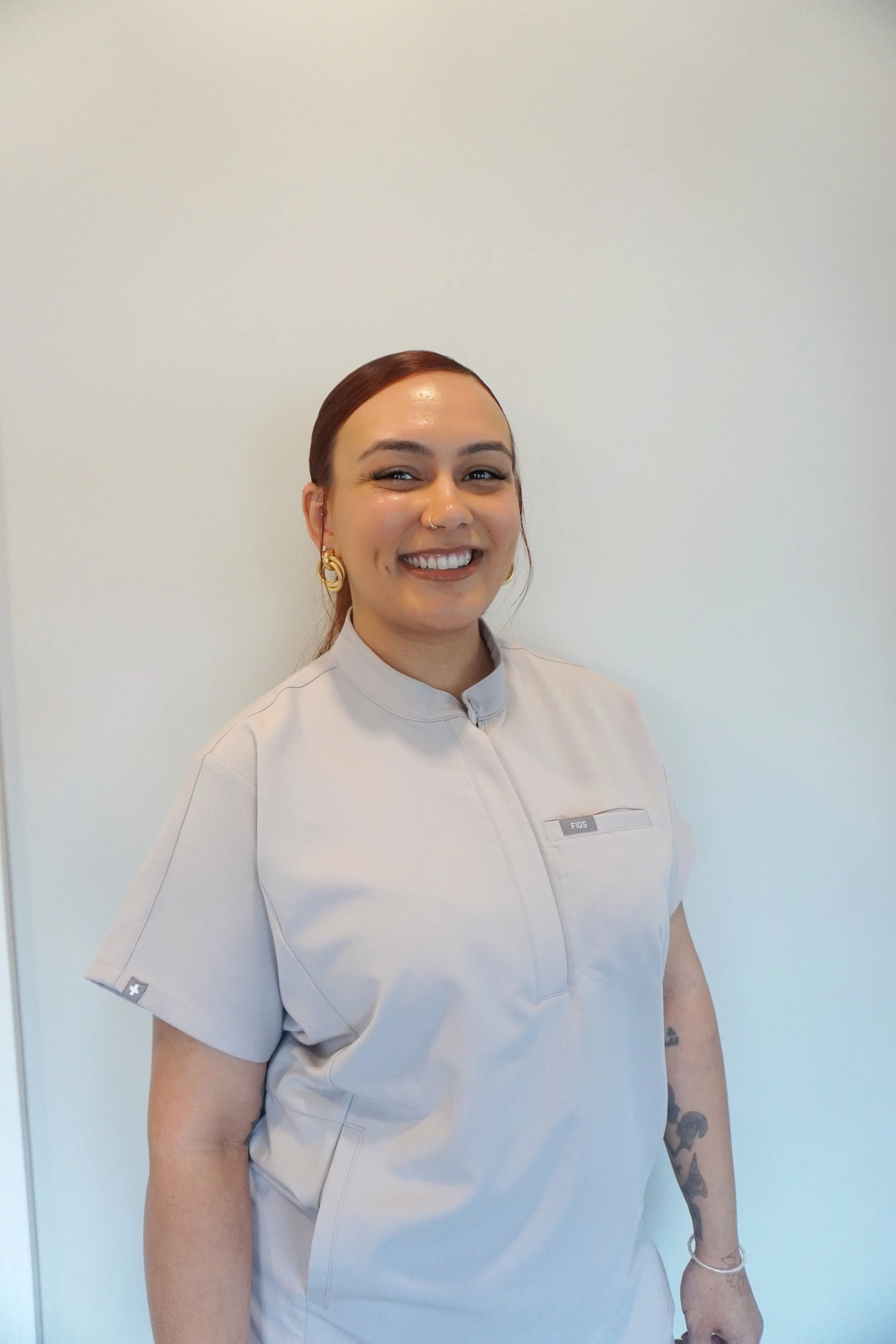 Justine wearing gold earrings, wearing a light gray medical uniform, standing against a plain light-colored wall.