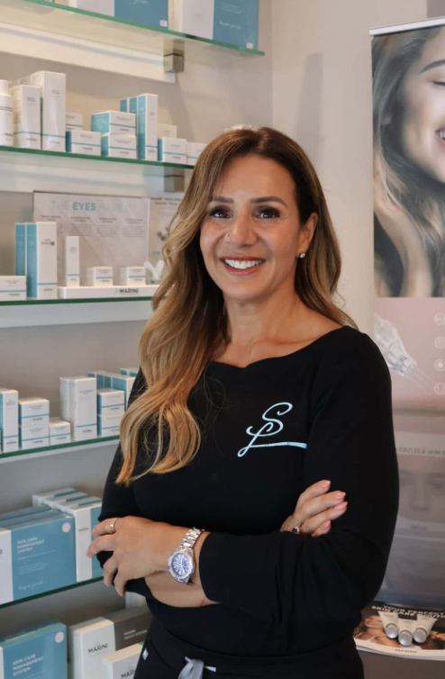 Kerri Sousa Owner of Spa Larimar smiling and standing in front of a retail display, wearing a black shirt with a light blue logo and a silver wristwatch.