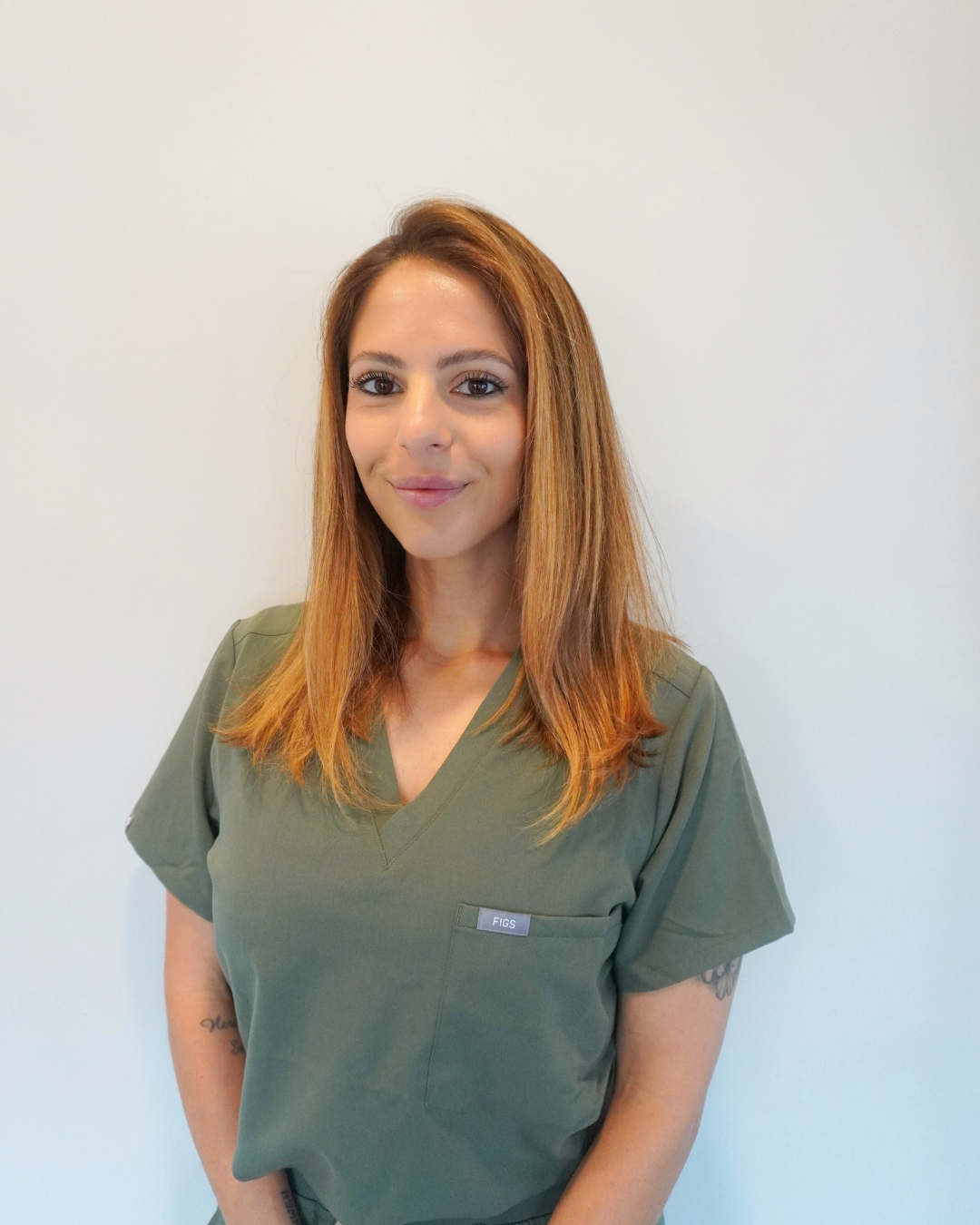 Sam with long light brown hair, wearing green scrubs, standing in front of a plain white wall with a slight smile.