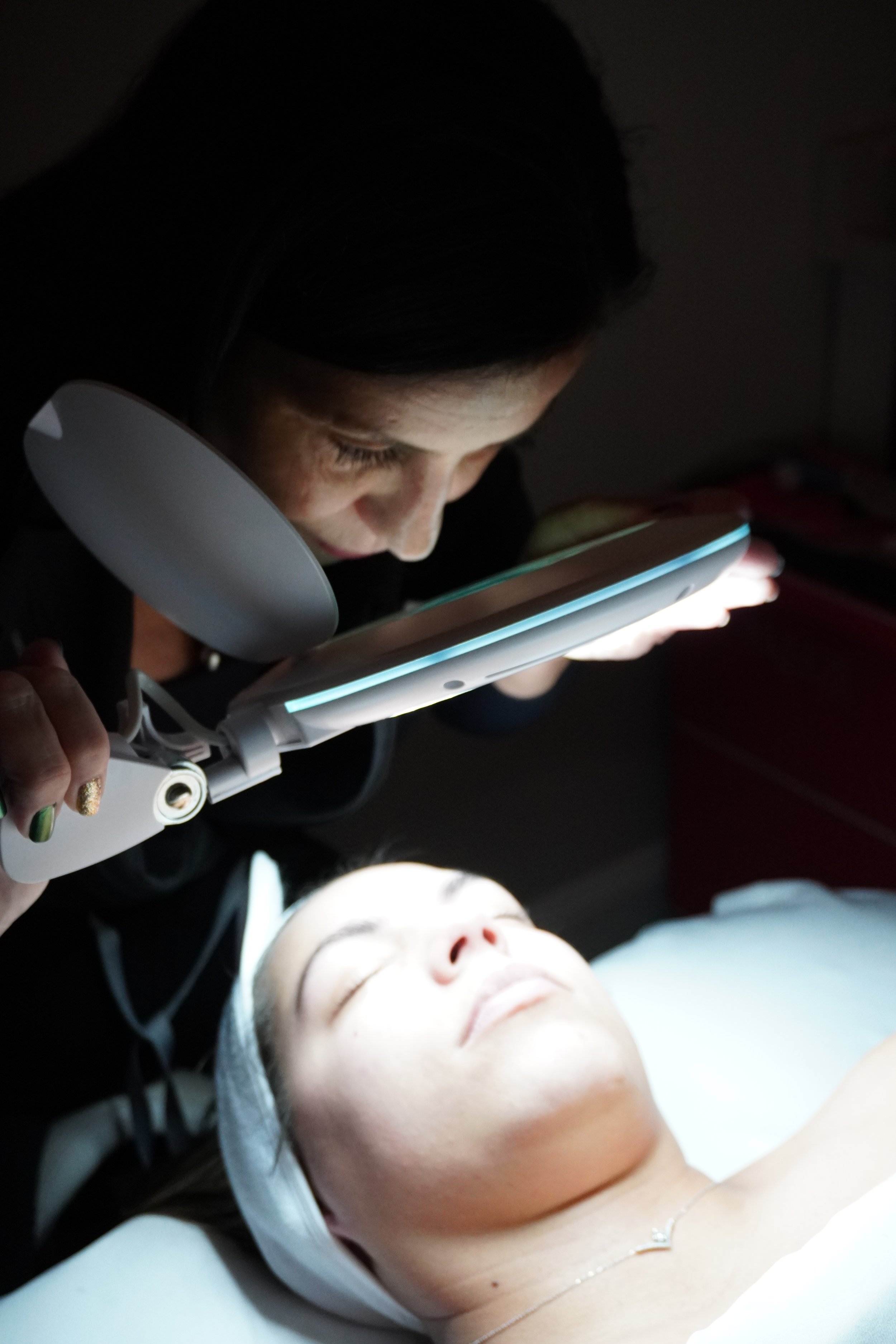 A woman undergoing a facial treatment with a device emitting light, while lying down with eyes closed, in a dimly lit spa or clinic setting.