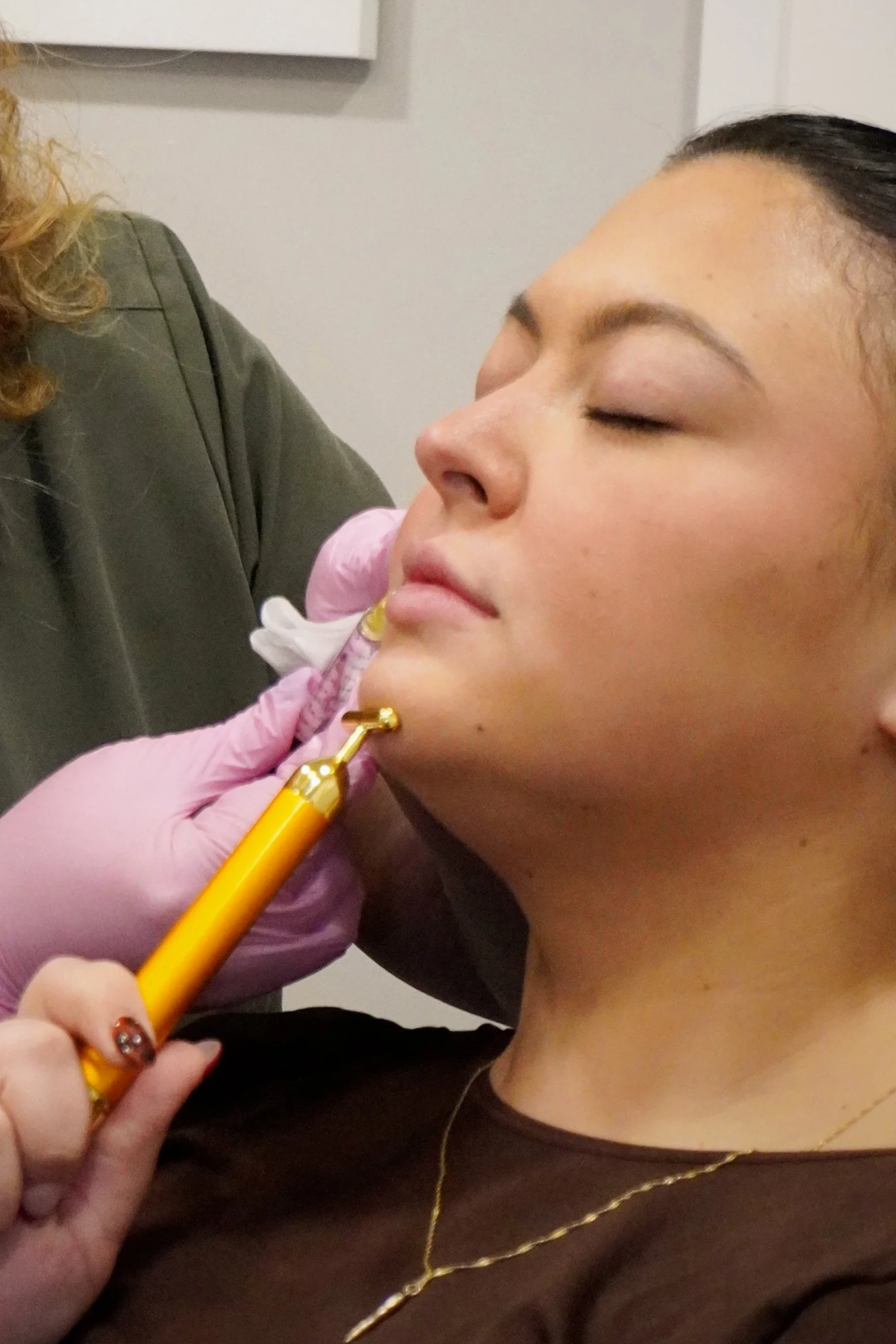 A woman receiving a cosmetic injection from a healthcare professional wearing pink gloves in a clinical setting.