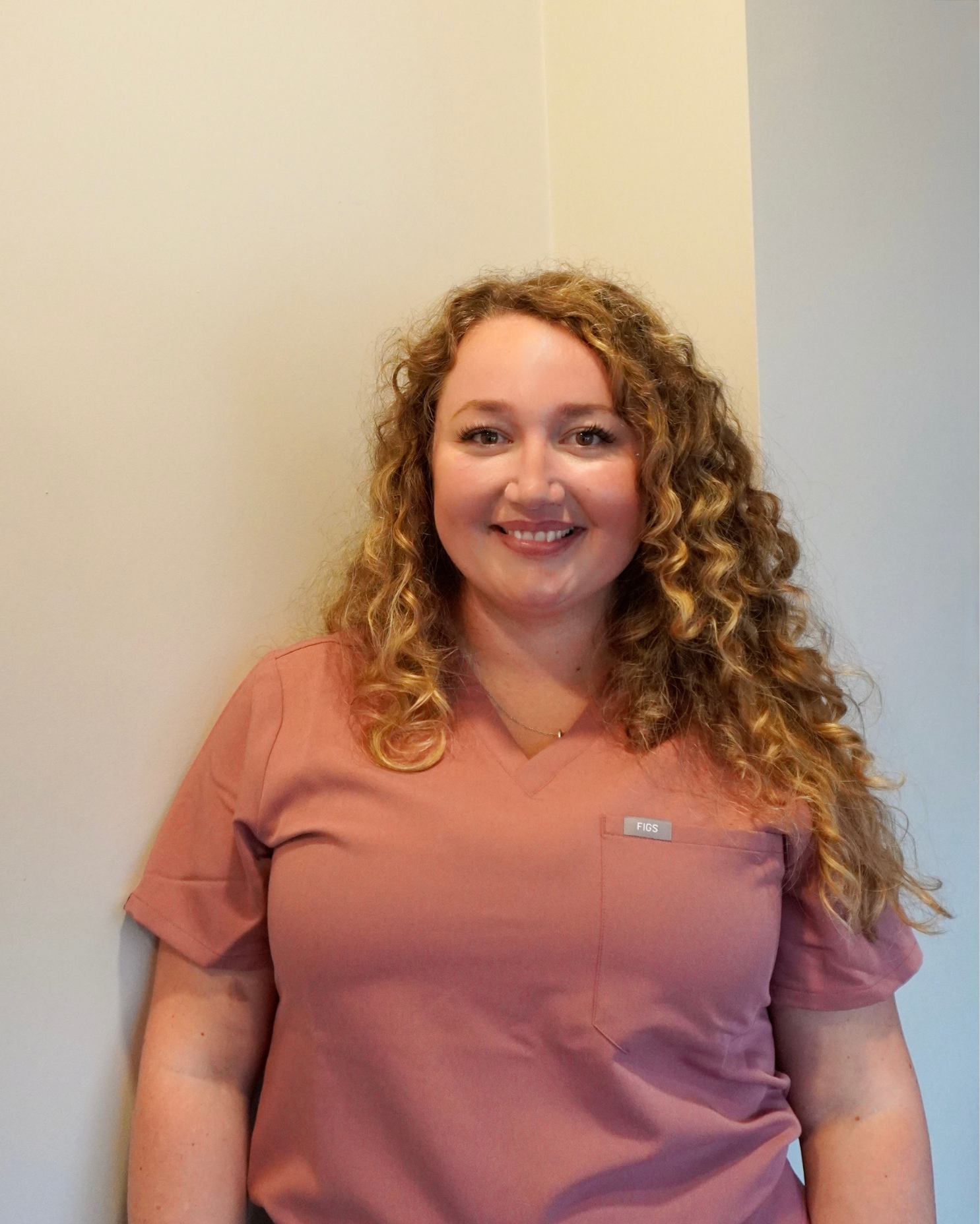 Ashley with curly blonde hair smiling, wearing a pink scrub top, standing against a plain light-colored wall.