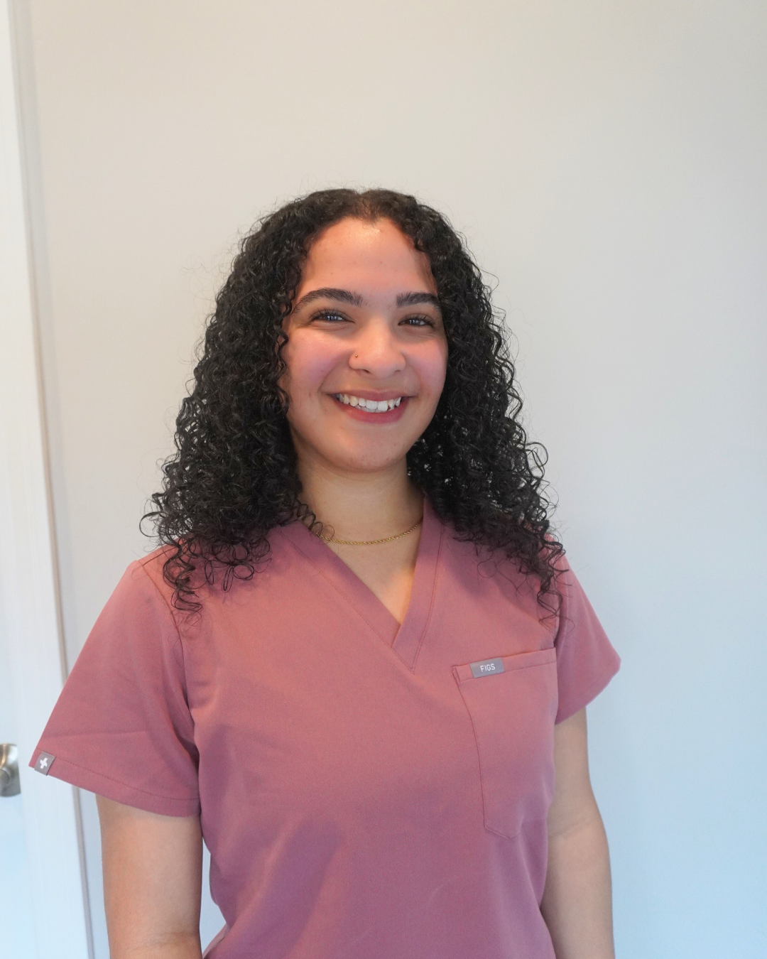 NyAsia wearing pink medical scrubs with curly black hair and a gold necklace, standing in front of a plain light-colored wall.