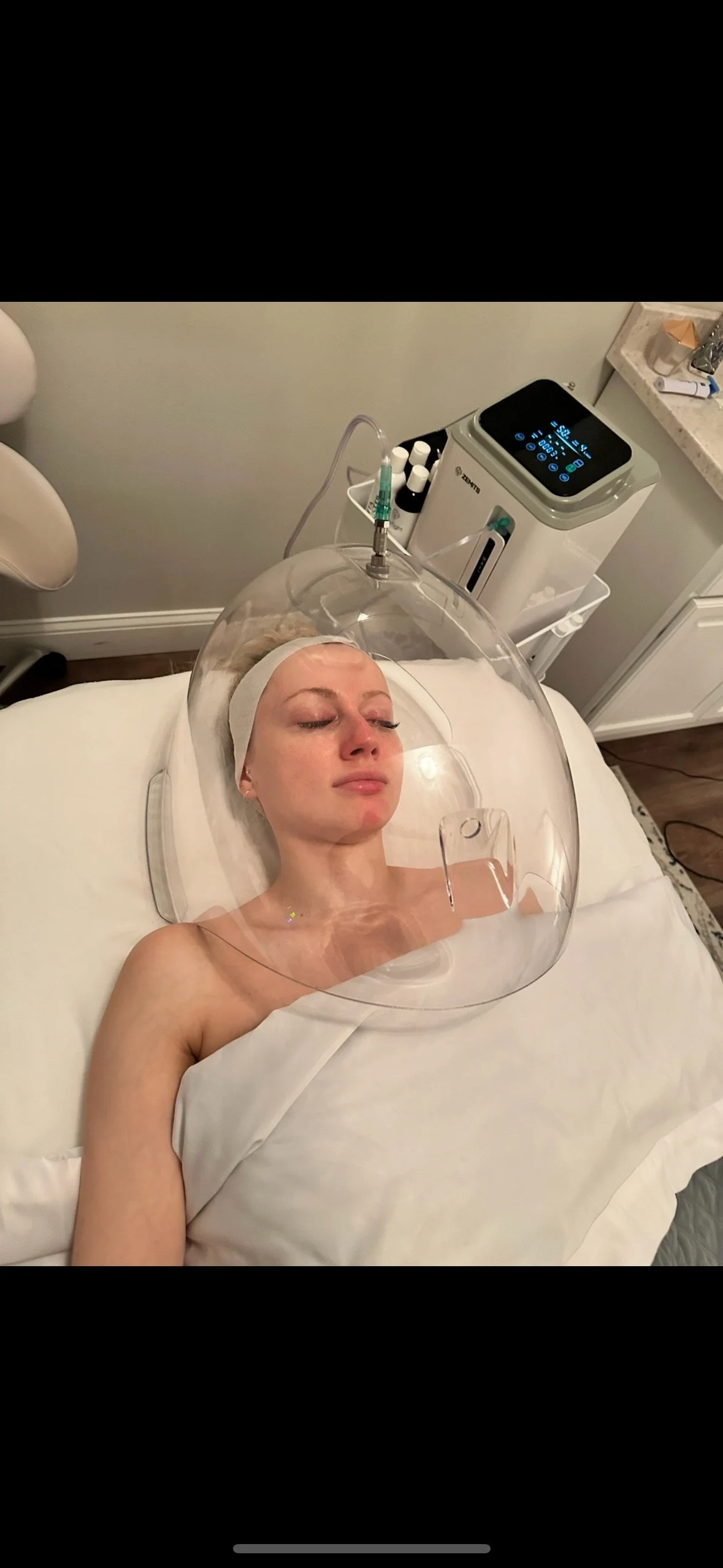 A woman lying on a hospital bed with her eyes closed, under a clear plastic dome, connected to a medical device, in a clinical setting.