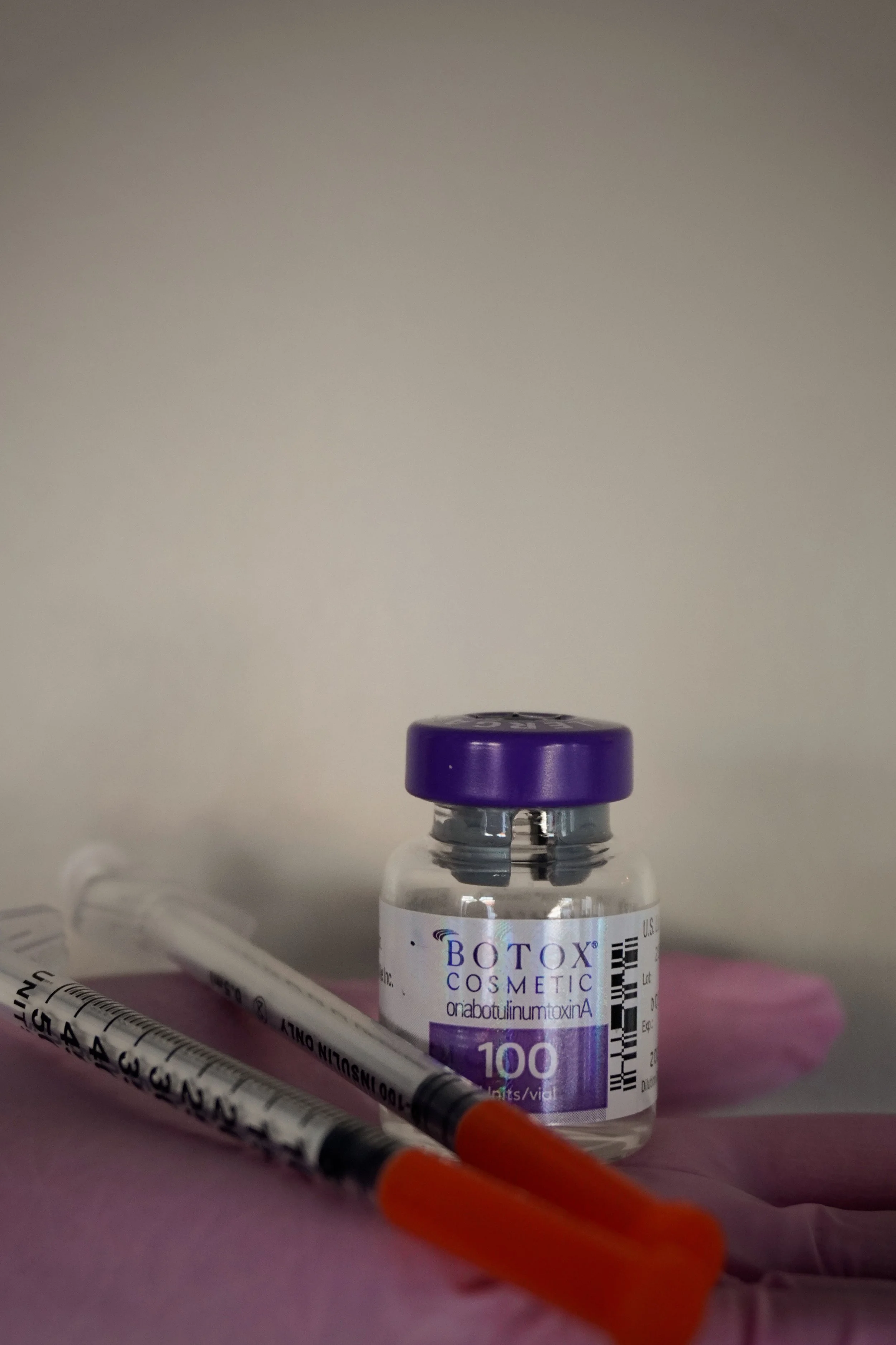 A small vial labeled 'Botox Cosmetic' with a purple cap, two syringes with orange needle covers, and a pink glove, set against a plain background.