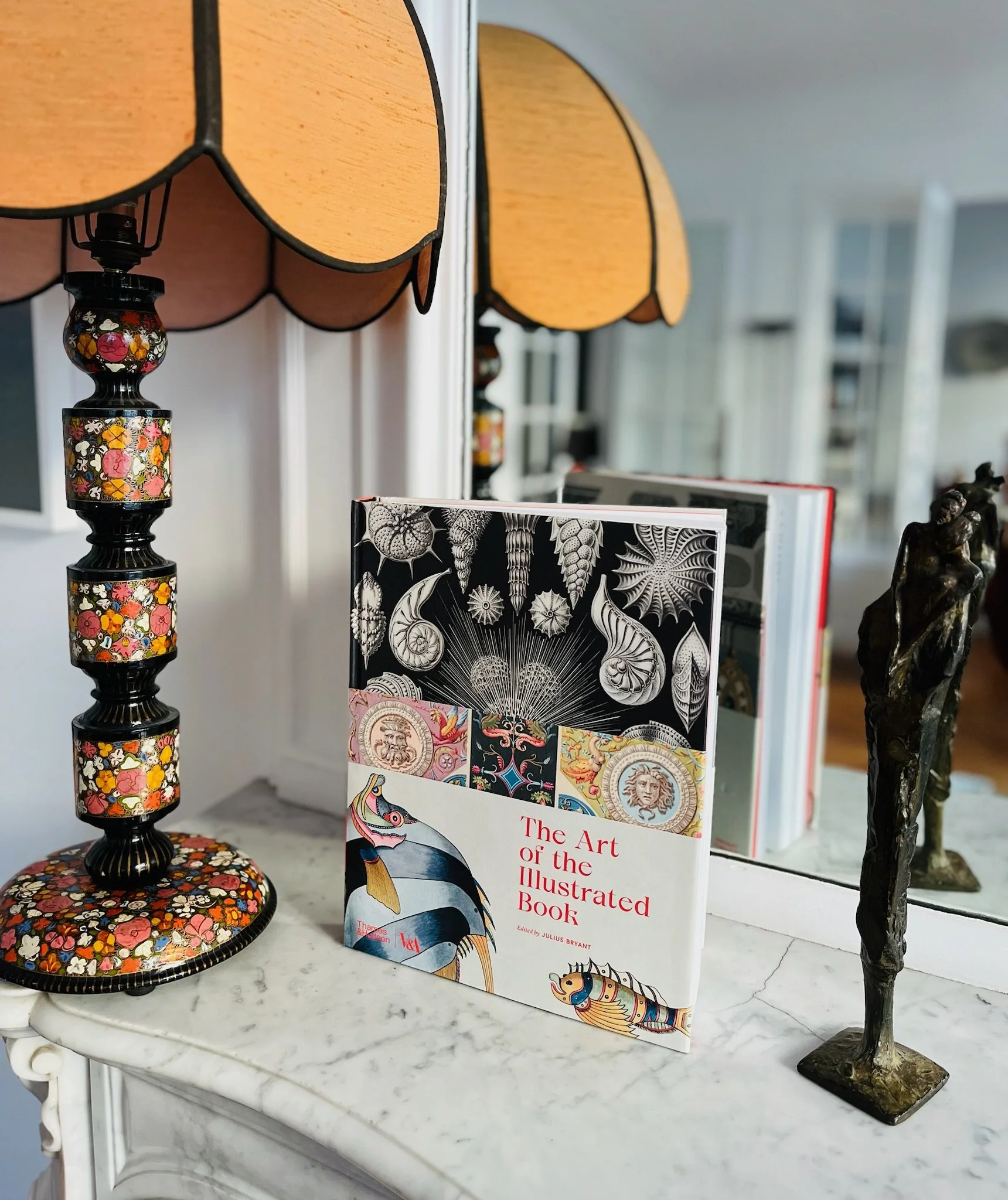 Livre intitulé "The Art of the Illustrated Book" posé sur une surface en marbre, à côté d'une lampe en mosaïque florale et d'une sculpture en bronze d'une figure humaine.