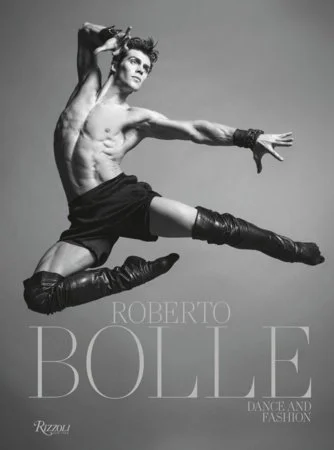 Un danseur masculin pose de manière expressive en portant des gants, des bottes hautes en cuir et un short noir, avec un fond gris et le texte 'Roberto Bolle, Dance and Fashion'