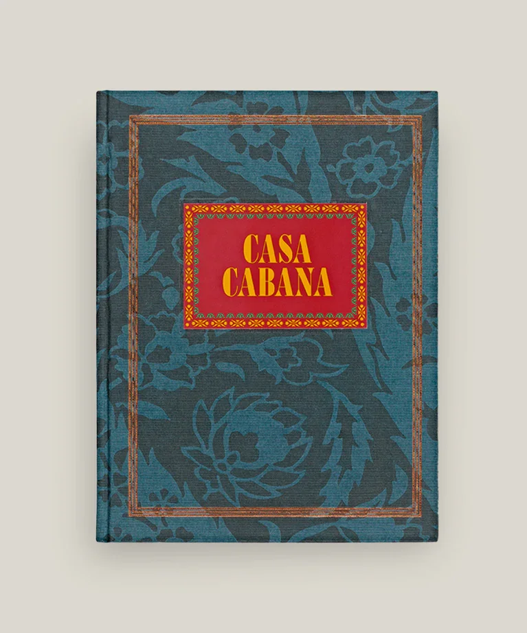 Livre intitulé 'Casa Cabana' avec une couverture bleue ornée de motifs floraux. Titre en lettres jaunes sur un fond rouge encadré de motifs rouges et jaunes.