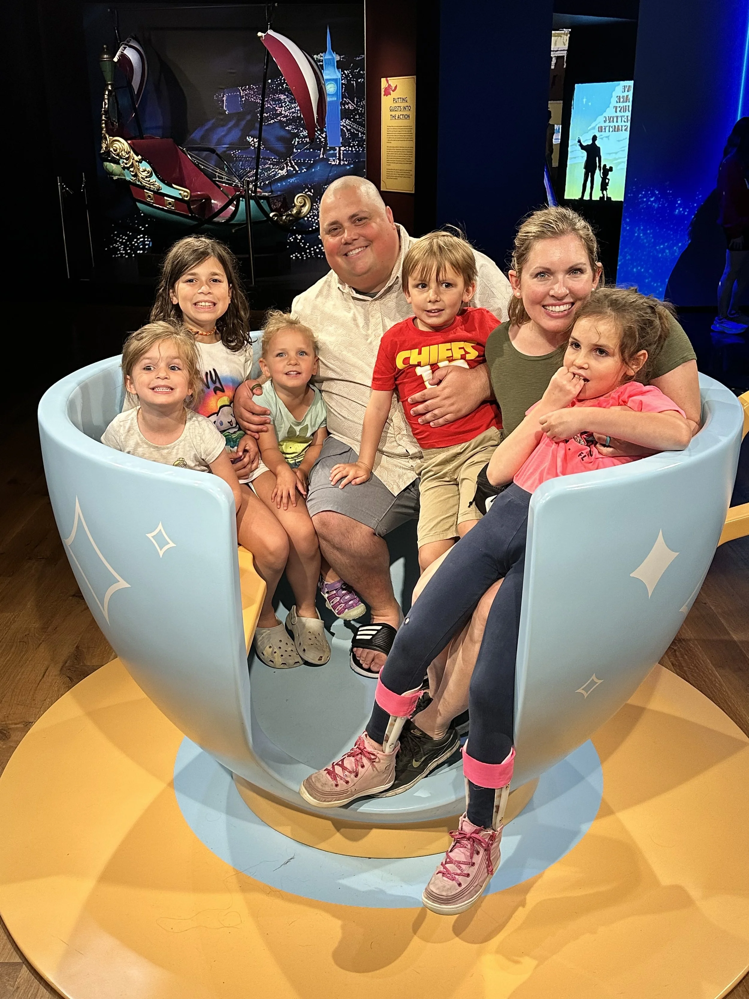 A family of seven seated inside a large, light blue teacup exhibit at a museum. The family includes three adults and four children smiling at the camera. In the background, there is a display of a boat and some signs.