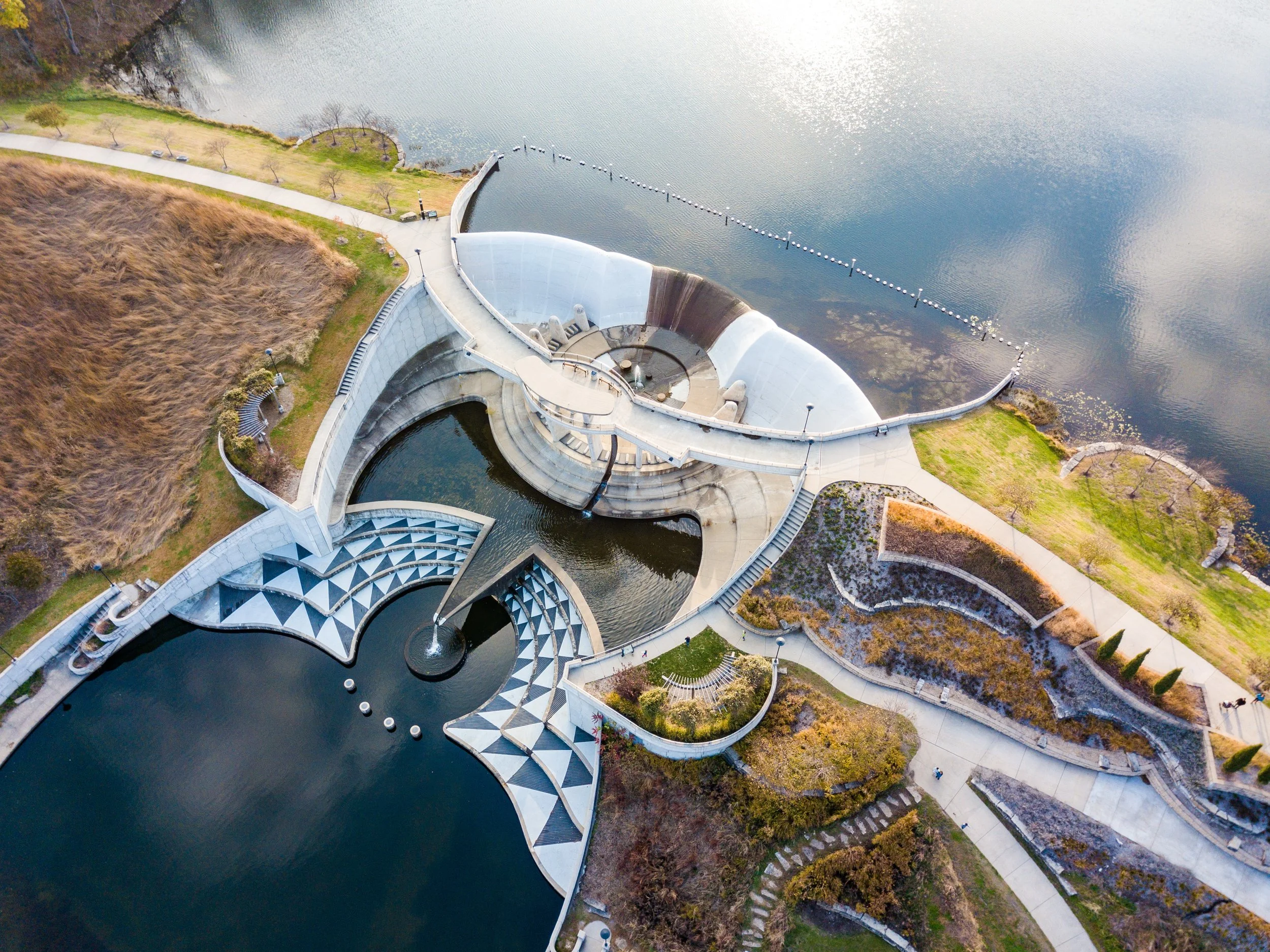 Aerial view of a park with a modern architectural structure featuring curved white walls and a fountain, adjacent to a body of water with a walking path and landscaped areas.