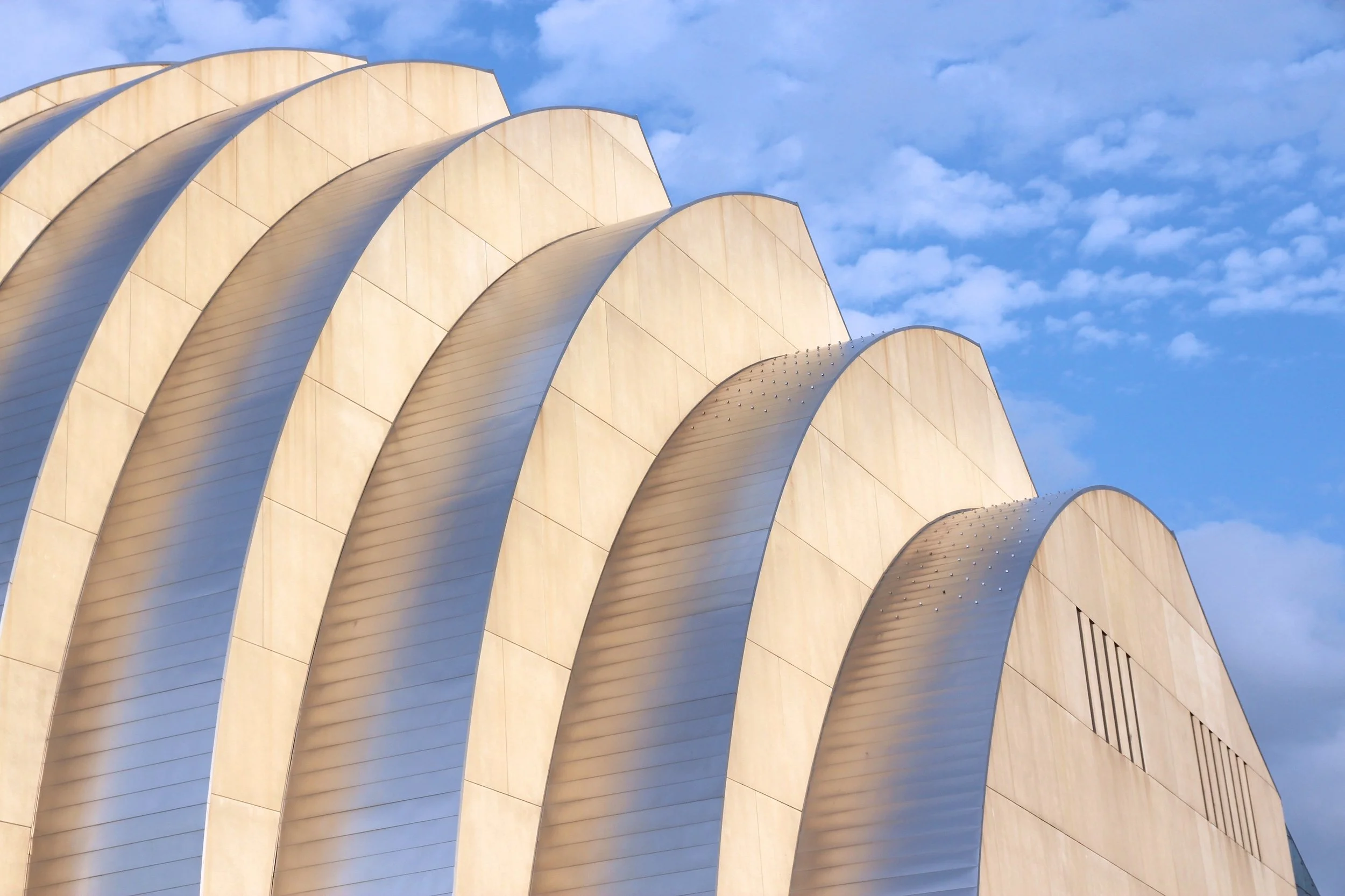 Modern building with curved, overlapping metallic panels and a beige exterior against a blue sky with scattered clouds.