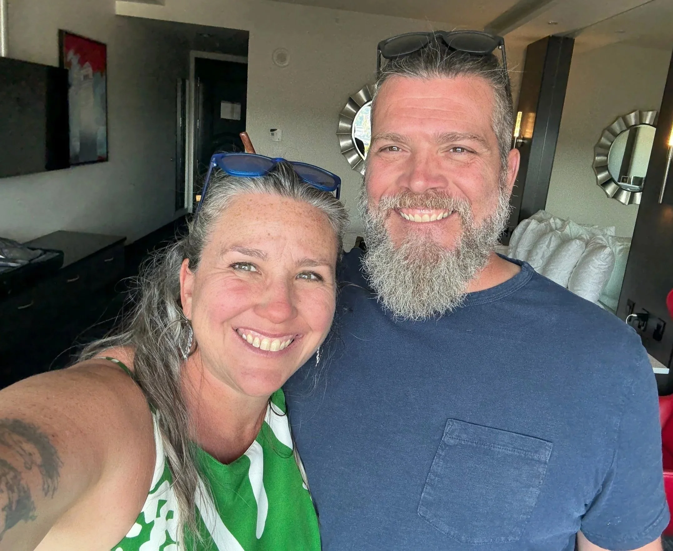 A smiling woman with tattooed arm and sunglasses on her head taking a selfie with a smiling man with a beard. They are inside a modern living room with decorative mirrors, a large bed, and a TV on the wall.