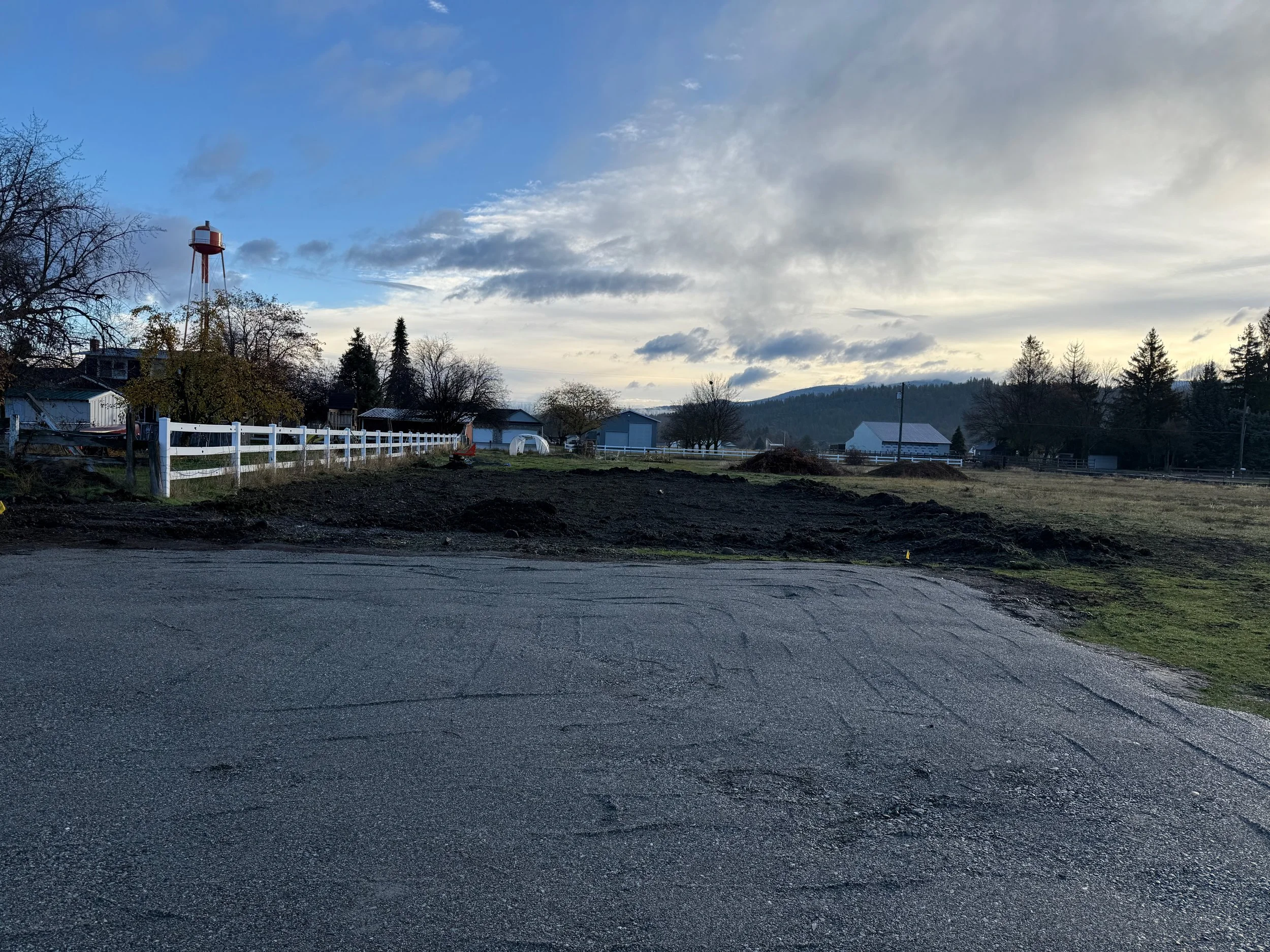 A rural scene with a blacktop road in the foreground, a white fence, trees, small houses, a water tower, and hills under a cloudy sky.
