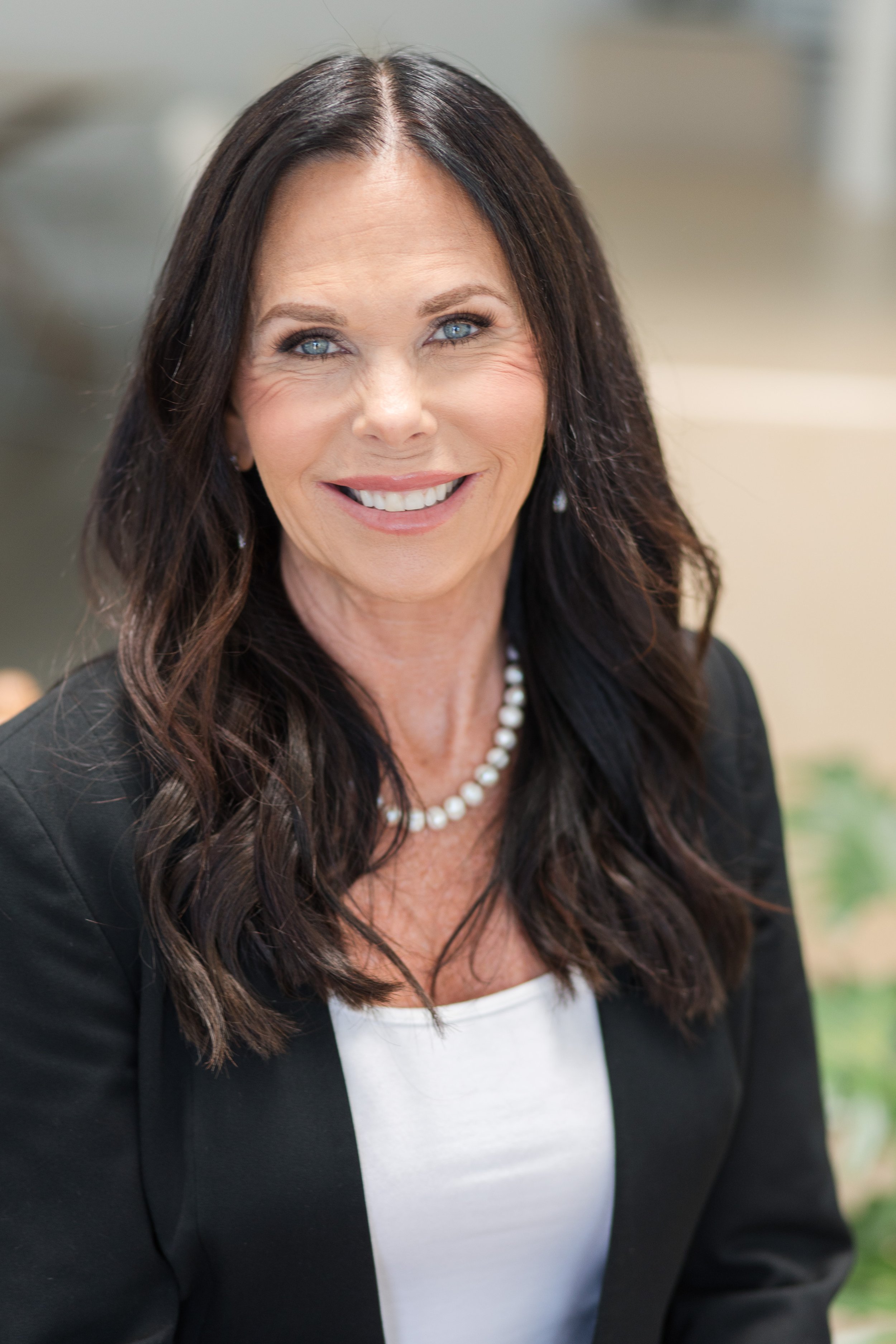 A smiling middle-aged woman with long dark hair, wearing a black blazer, a pearl necklace, and a white top. Presented in a professional setting.