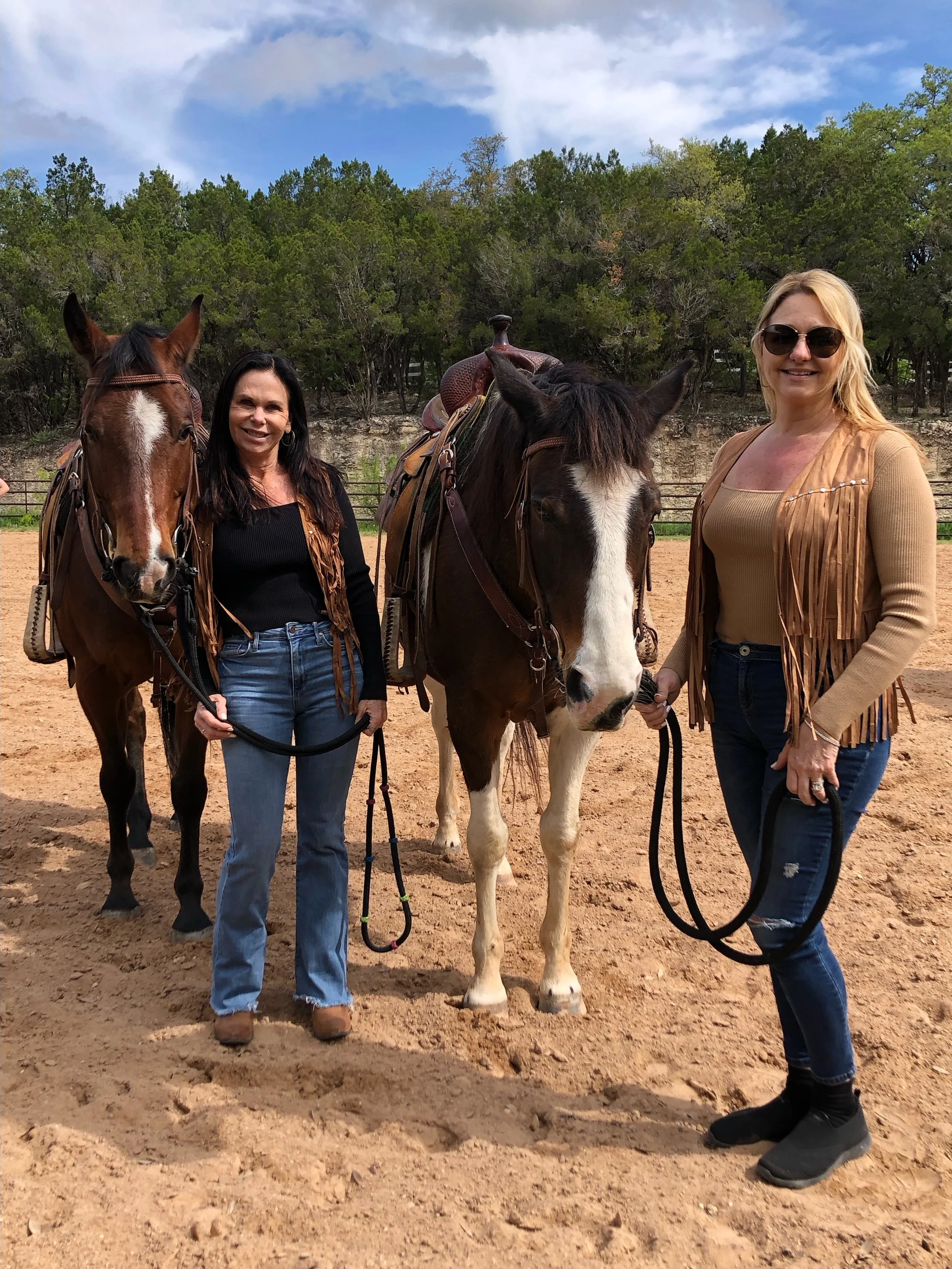 Two women standing on a dirt field holding the reins of two horses, with a forest and blue sky in the background.