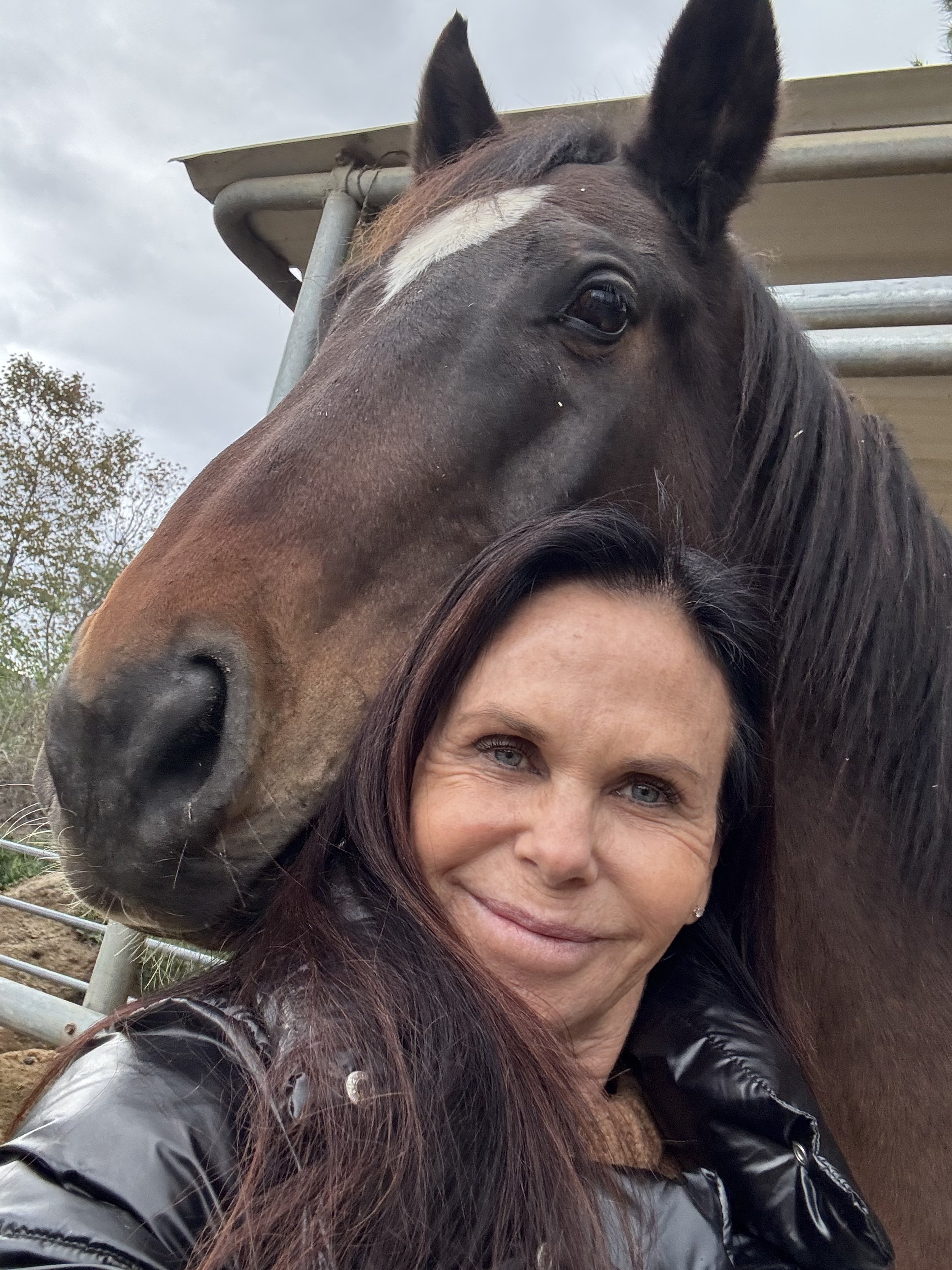 A woman with long dark hair taking a selfie with a large brown horse, both close to the camera, outdoors during cloudy weather.