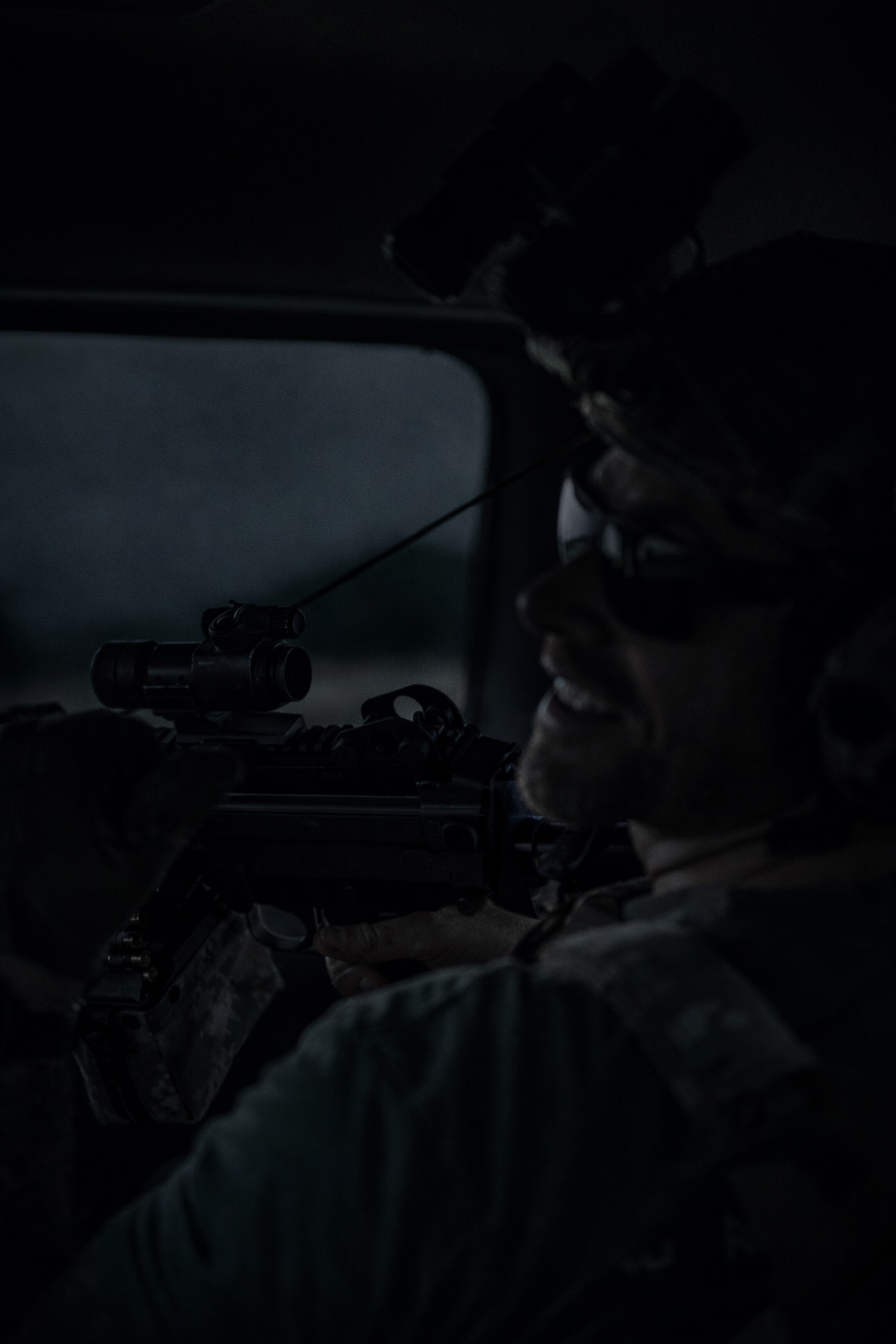 A soldier in camouflage uniform and helmet aiming a scoped rifle inside a dark vehicle.