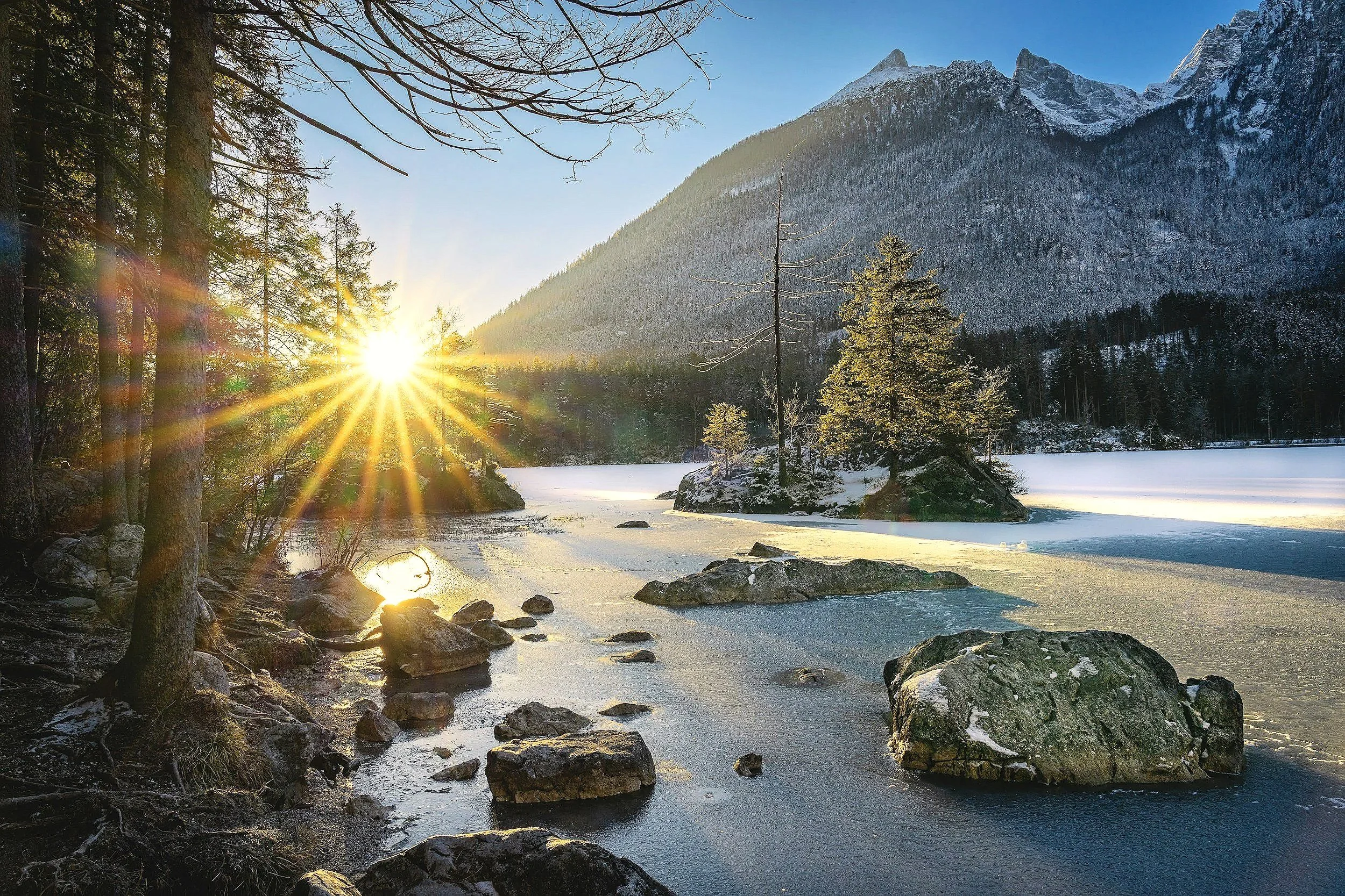 Sunrise over a snow-covered mountain landscape with a frozen river, rocks, and trees in a winter forest.