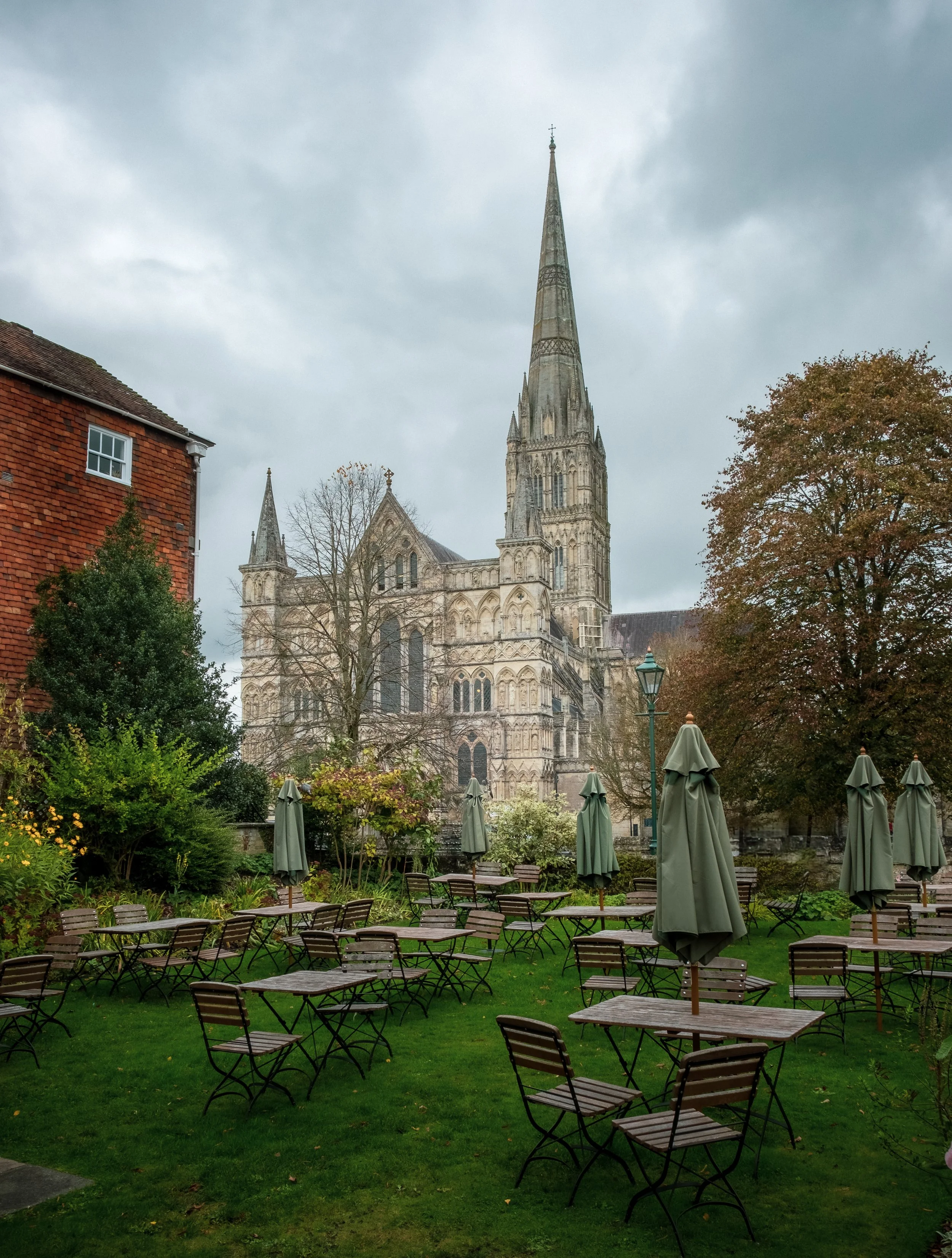 Salisbury Cathedral and an outdoor garden with wooden tables and chairs, umbrellas, trees, and a Gothic-style cathedral in the background under a cloudy sky.