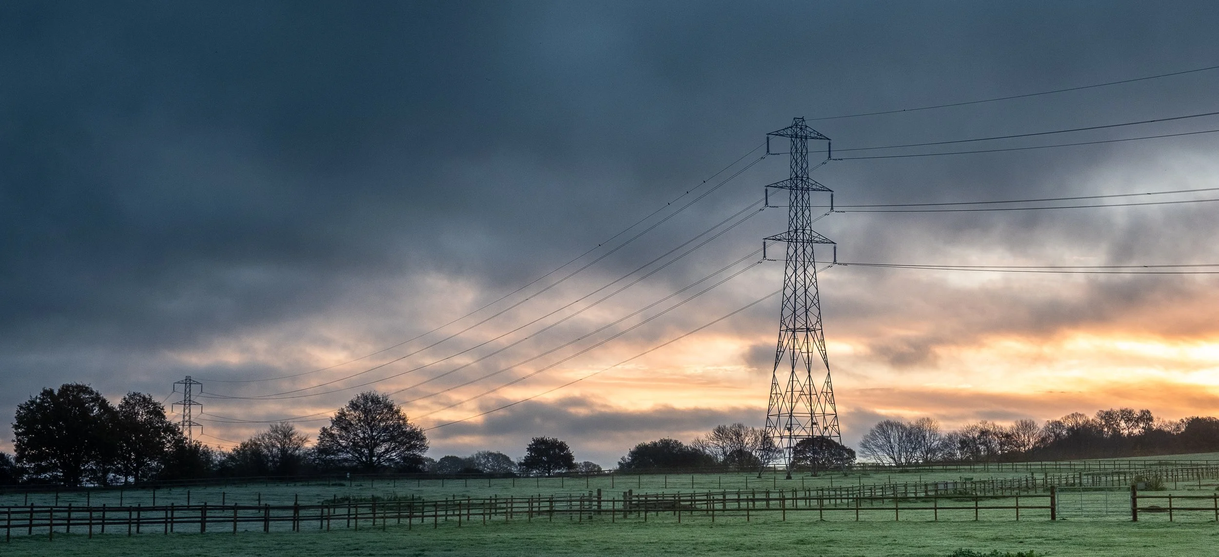 A rural landscape at sunrise with a large electrical transmission tower, power lines, a fence, and trees in the background under a cloudy sky.