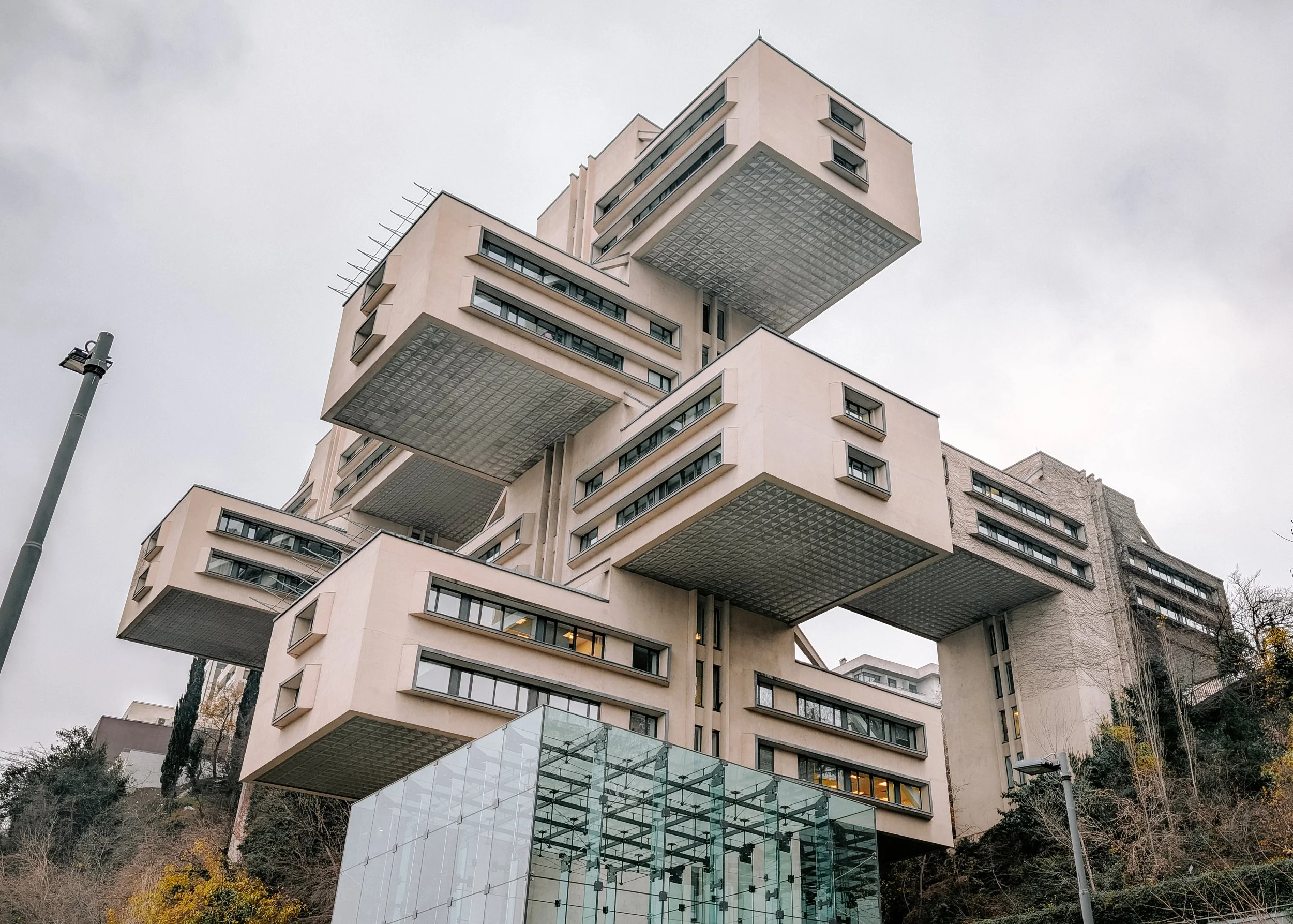A modern multi-story building with protruding cube-like structures and glass accents, set against an overcast sky. The building has a unique architectural design with cantilevered sections and appears to be built on a hill with some trees and streetl