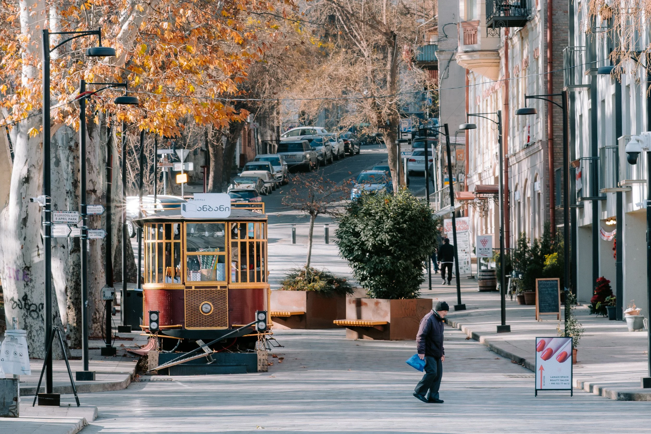Empty city street with a vintage tram train stopped on the sidewalk, surrounded by trees with autumn-colored leaves and storefronts with awnings. A person walks across the street carrying a blue bag.