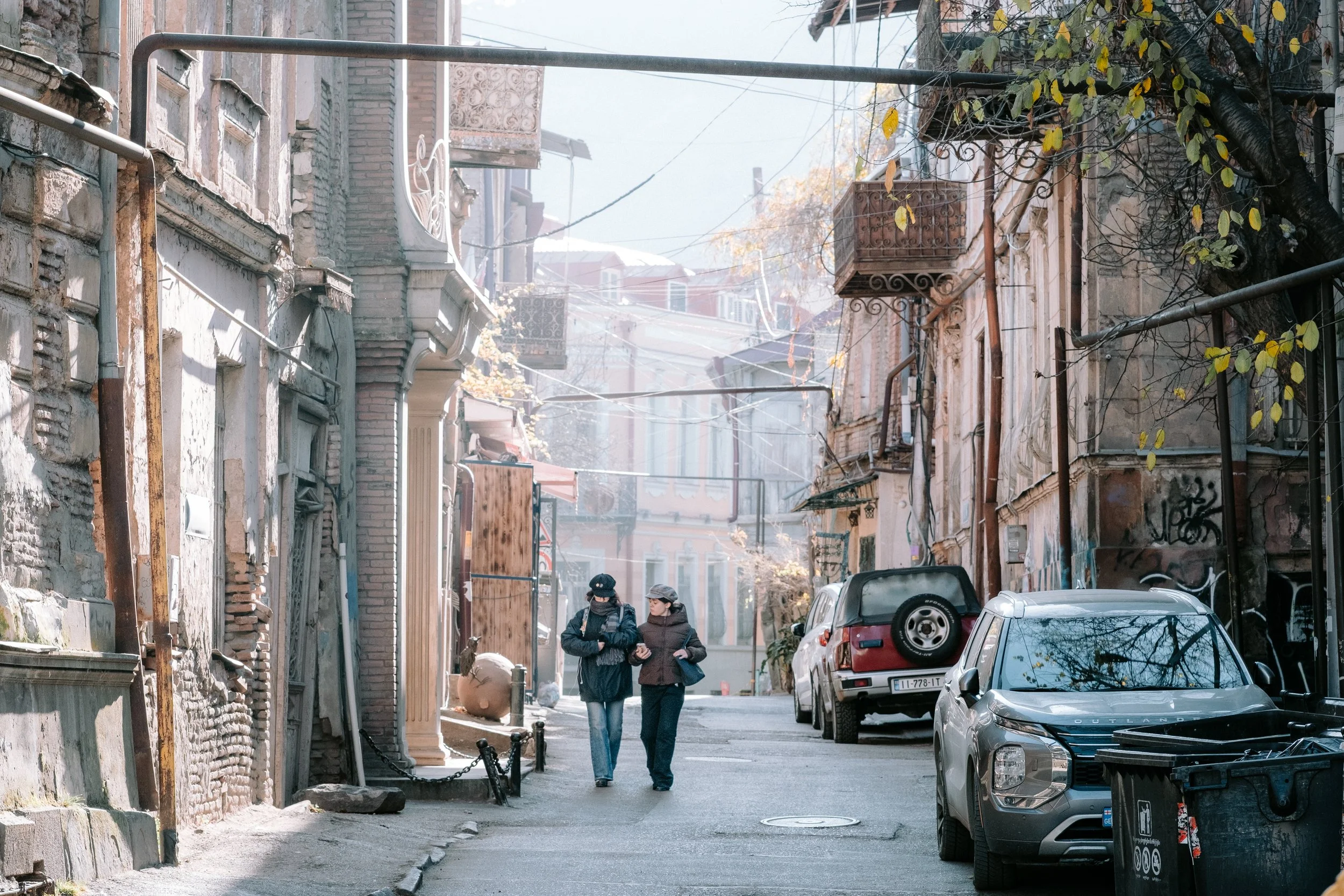 Two women walking and talking on a narrow street lined with old buildings, parked cars, and graffiti. One woman is wearing a black mask and the other a gray hat.