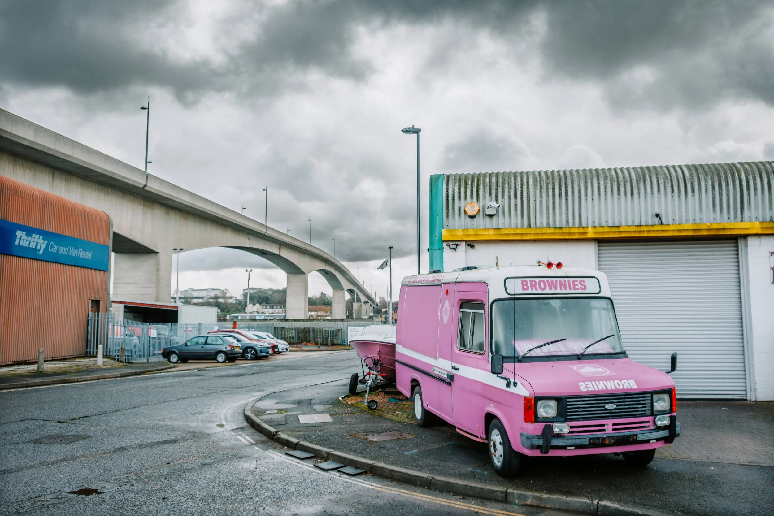 A pink ice cream truck with a sign that says 'BROWNIES' parked outside a building with a gray metal roof. There are several parked cars near the building, and a bridge can be seen in the background under a cloudy sky.