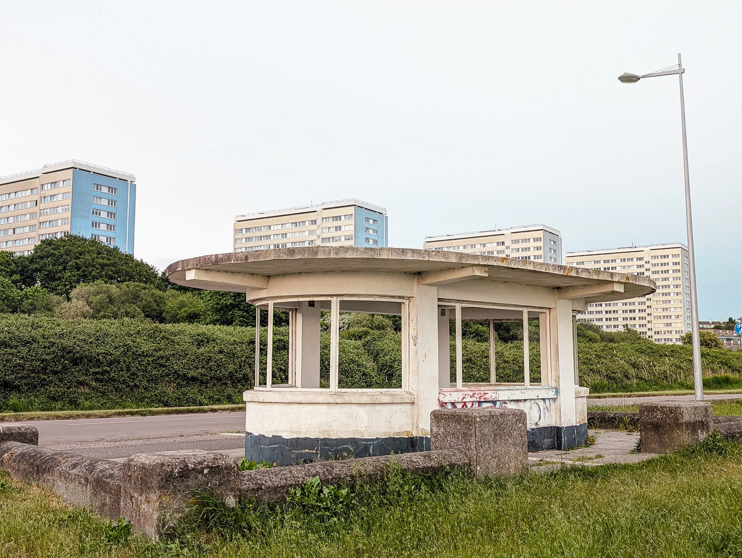 An old, small, white bus stop with large windows and graffiti on the wall, surrounded by grass and concrete blocks, with tall apartment buildings and a streetlamp in the background.