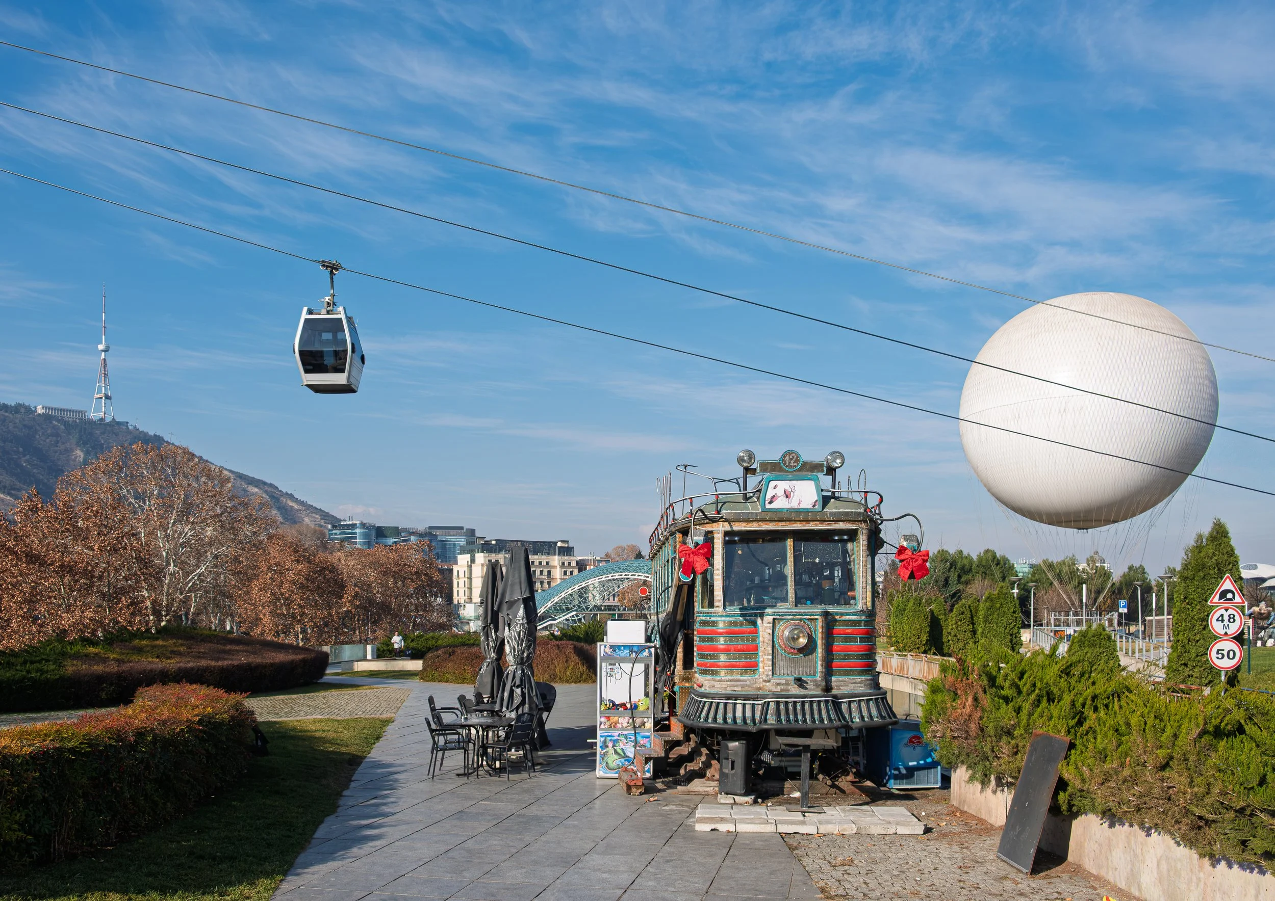 An outdoor scene with a gondola lift, a large spherical structure, and an old train carriage decorated with red ribbons, situated along a paved pathway with chairs and umbrellas under a partly cloudy sky.