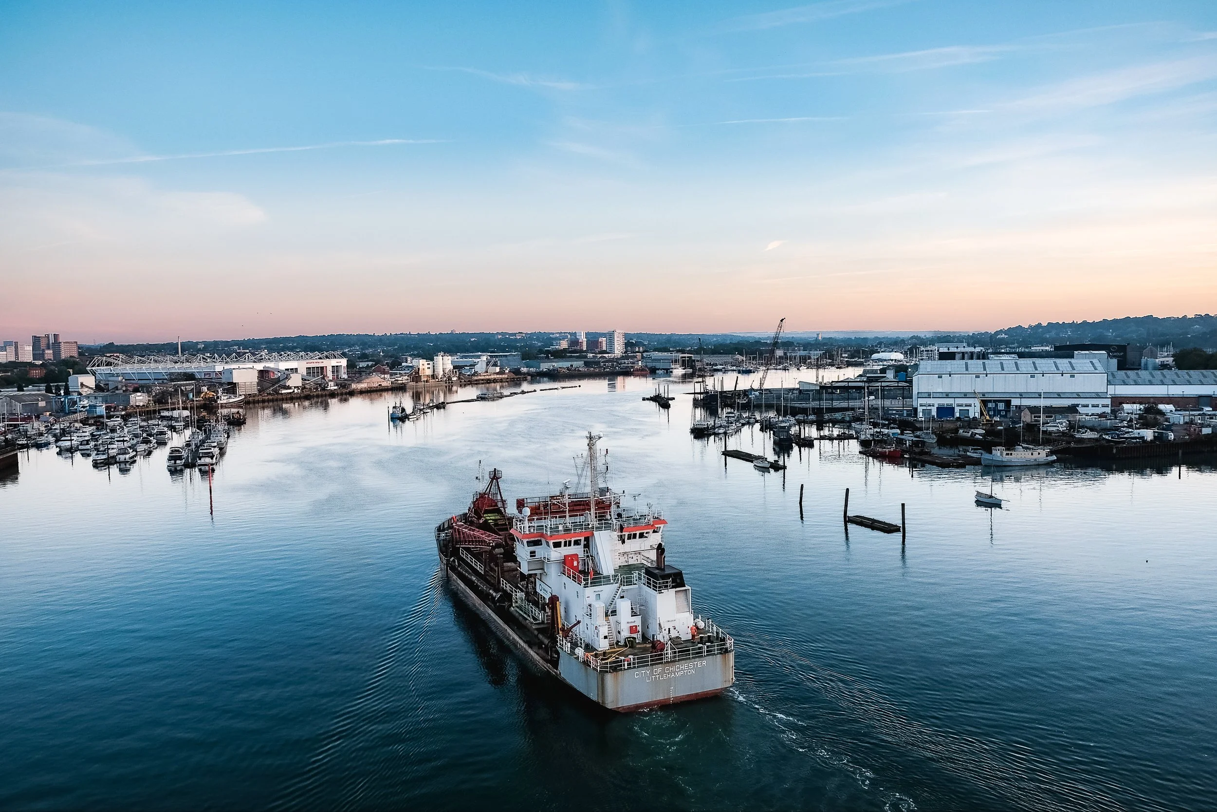 Aerial view of a harbor with a cargo ship moving through calm water, docked boats, and industrial buildings in the background at sunrise.