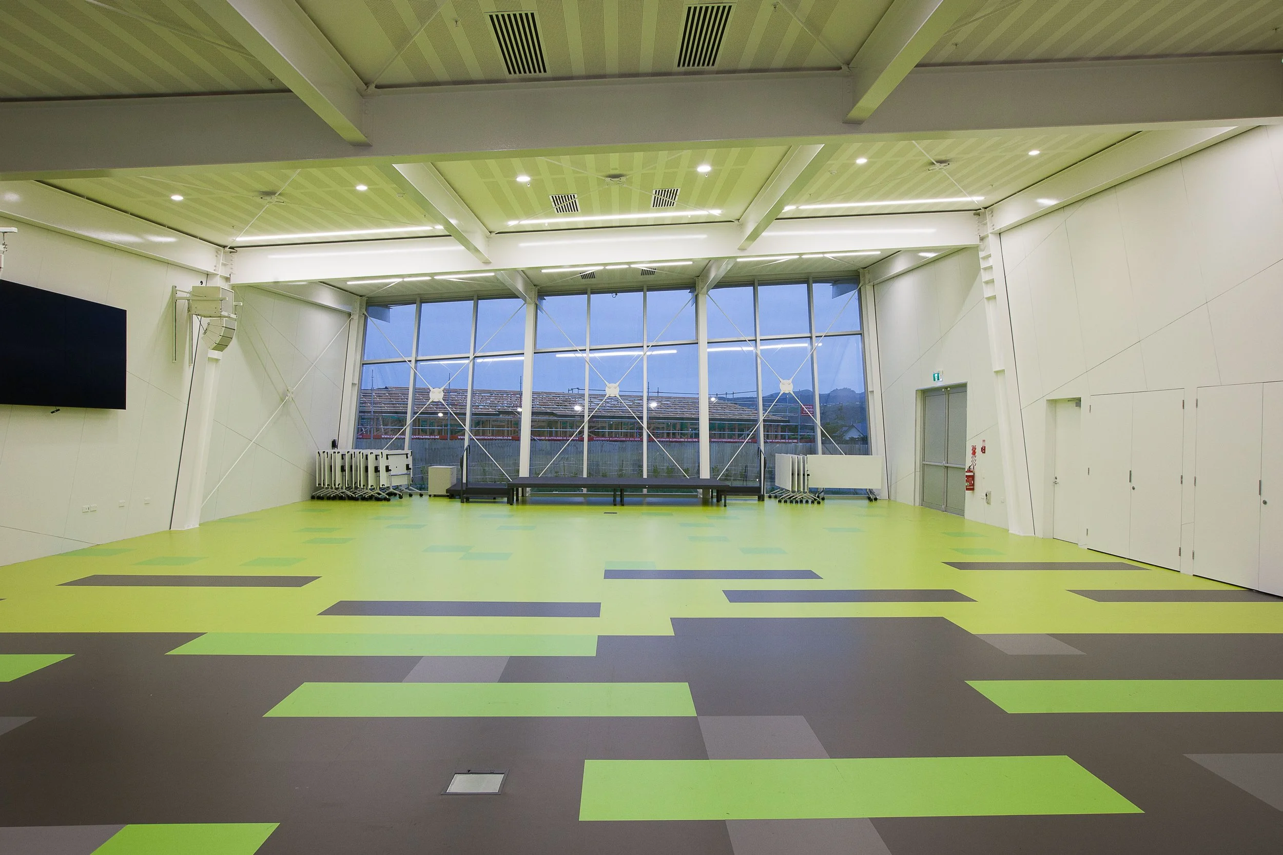 An empty indoor gym or multi-purpose room with bright green and gray patterned flooring, white walls, large windows showing an outdoor view, and ceiling lights.