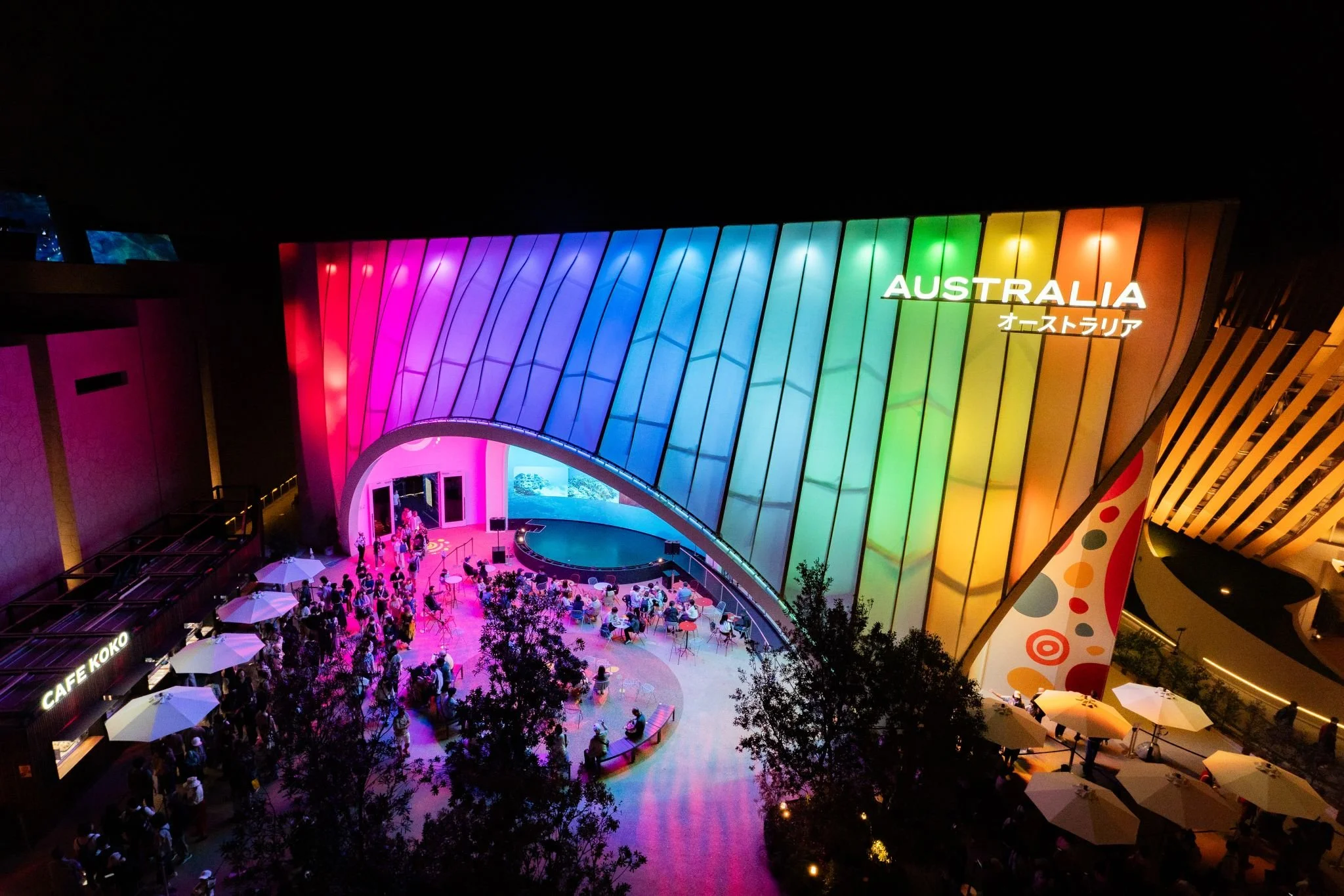 Night view of an illuminated building with a large rainbow-colored glass facade and the word 'Australia' on it, with people gathered outside under umbrellas.