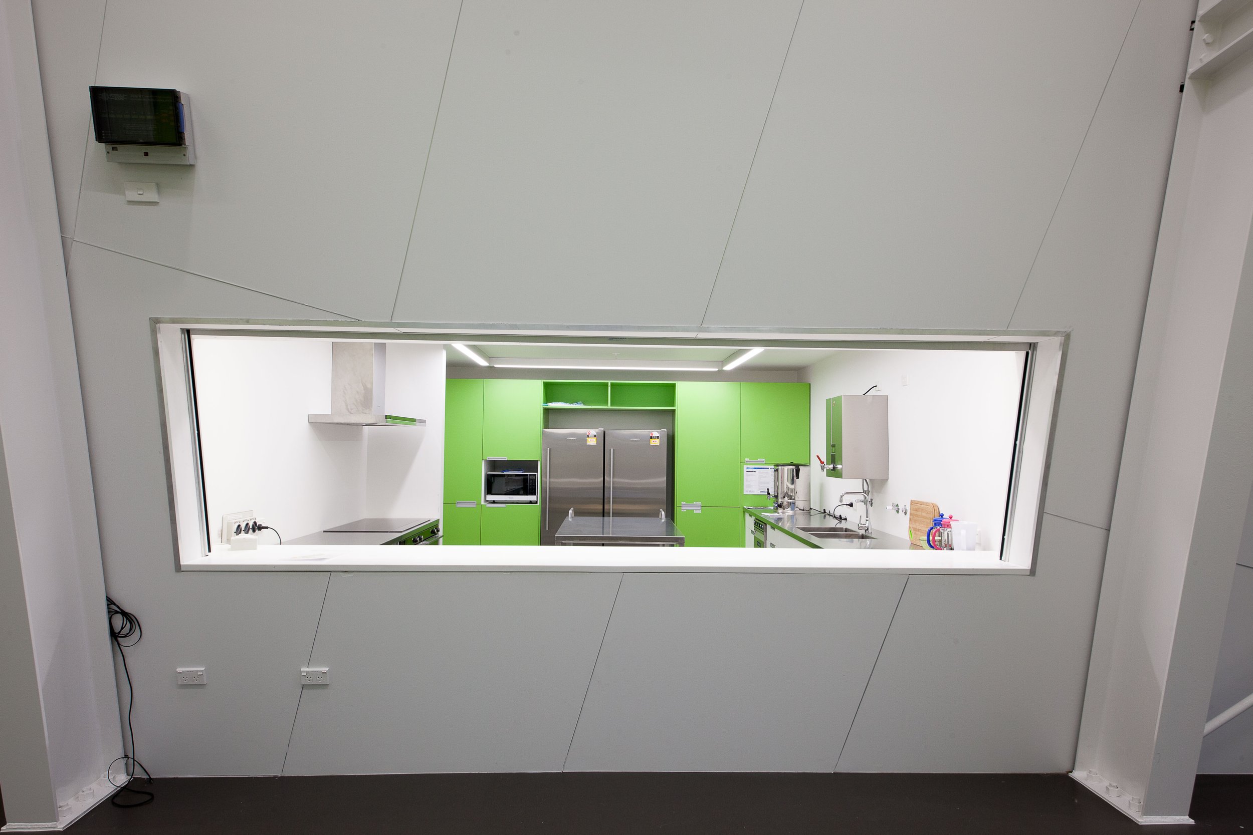Snapshot of a modern kitchen visible through a rectangular opening in a white wall, with green cabinets, stainless steel appliances, and a white countertop.