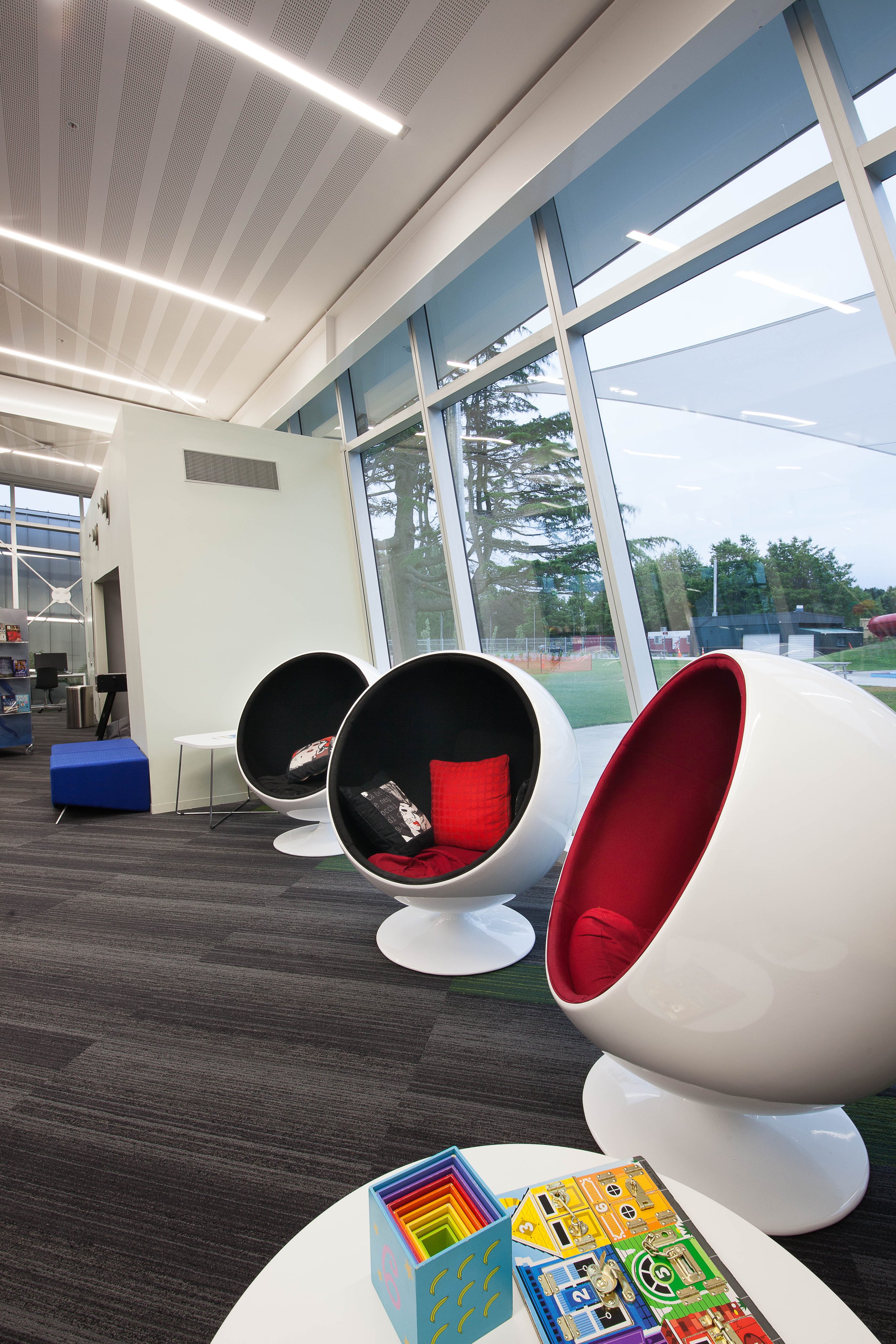 Modern lounge area with white spherical chairs that have red and black cushions, a small white table, and a colorful rainbow-shaped toy set on a white table. Large windows reveal a view of trees and outdoor space.