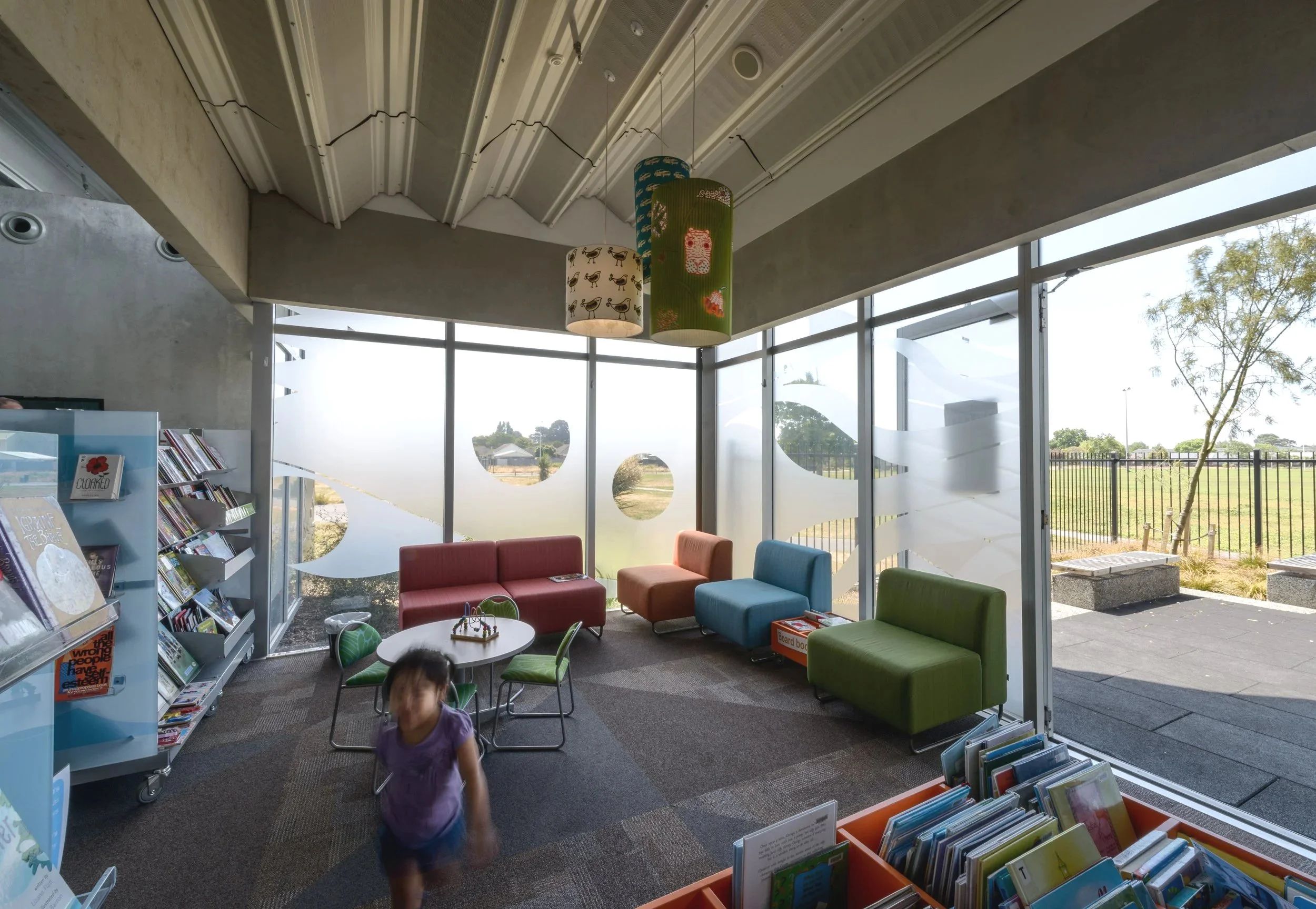 Children's reading area with colorful chairs, a small round table, bookshelves, large glass windows with decorative frosted patterns, and a young girl in purple shirt walking in a library or community center.