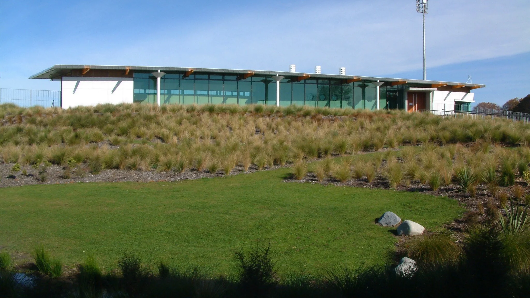 Modern building with large glass windows on a hill, surrounded by grass and ornamental grasses, with a clear sky in the background.