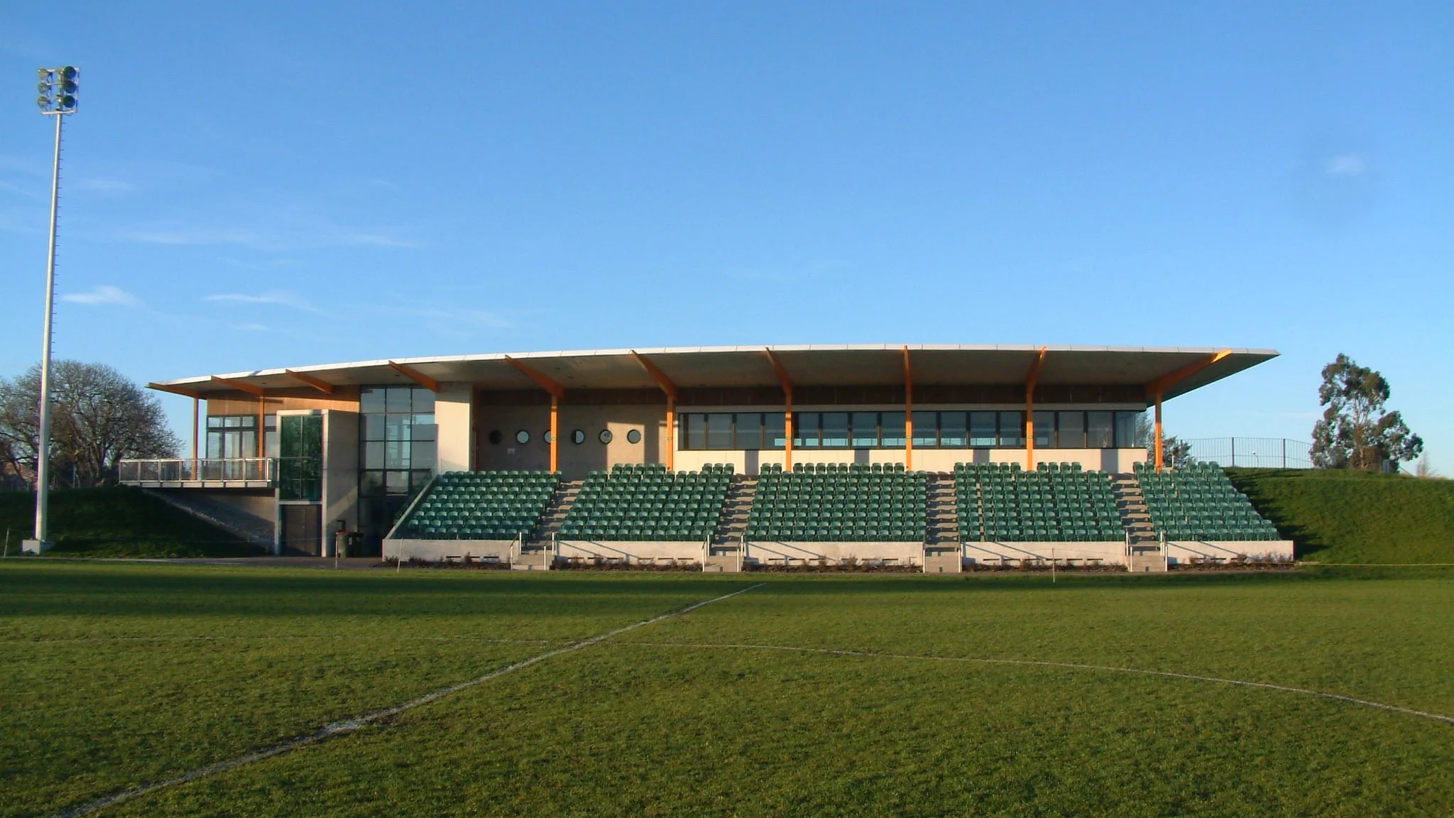 Empty sports stadium with green seating, a grass field, and a modern building with large glass windows and a curved roof in the background under a clear blue sky.