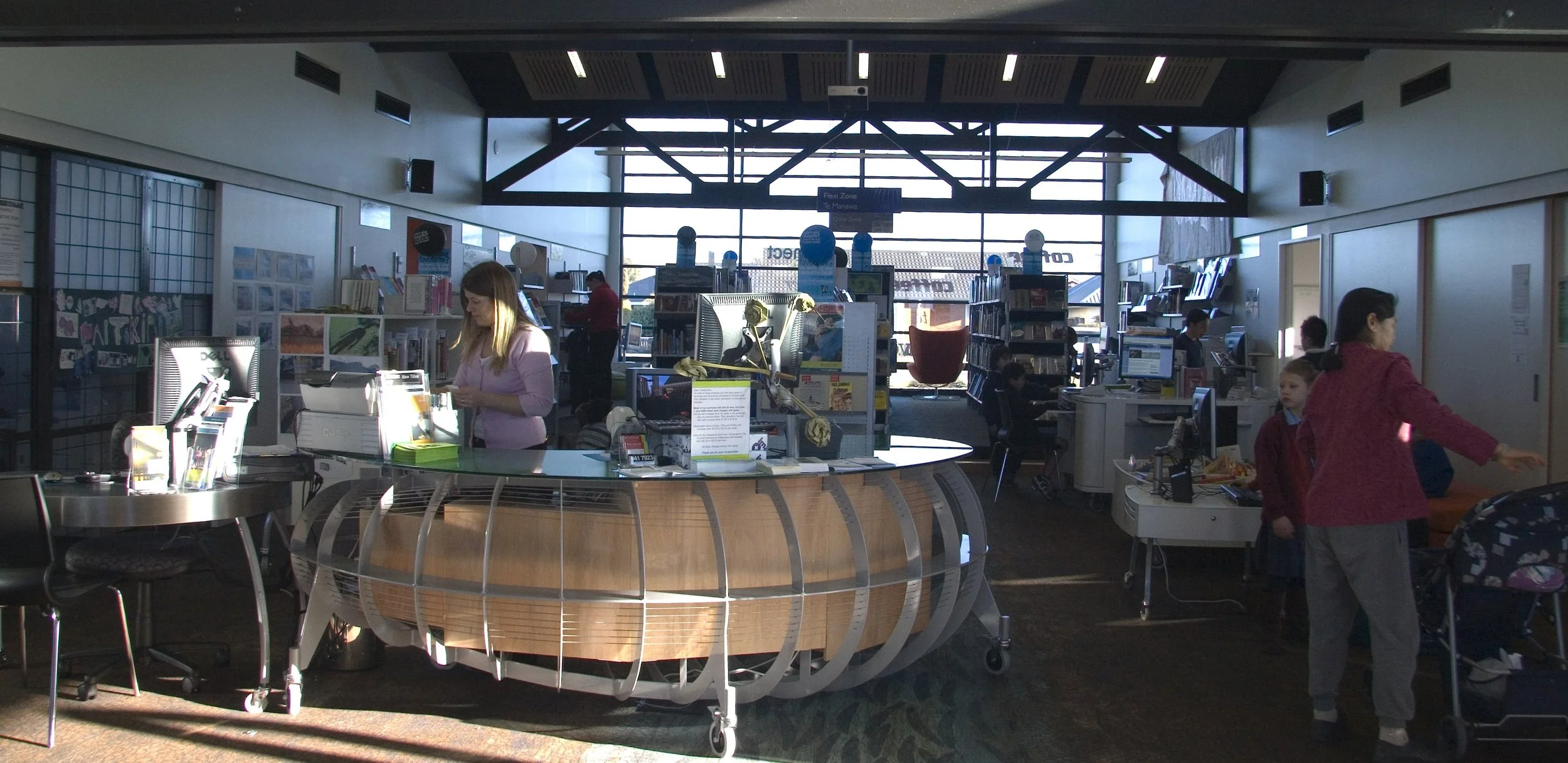 Inside a library with a service desk, shelves of books, and several children and adults. Sunlight streams through large windows at the back.