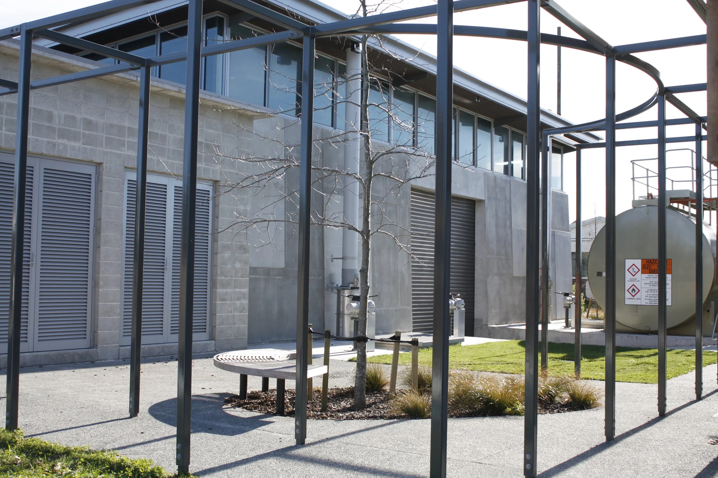 Modern building with gray concrete walls and large windows, a leafless tree, and a metal structure frame surrounding a small landscaped area with grass, a park bench, and a large industrial tank with safety signs.