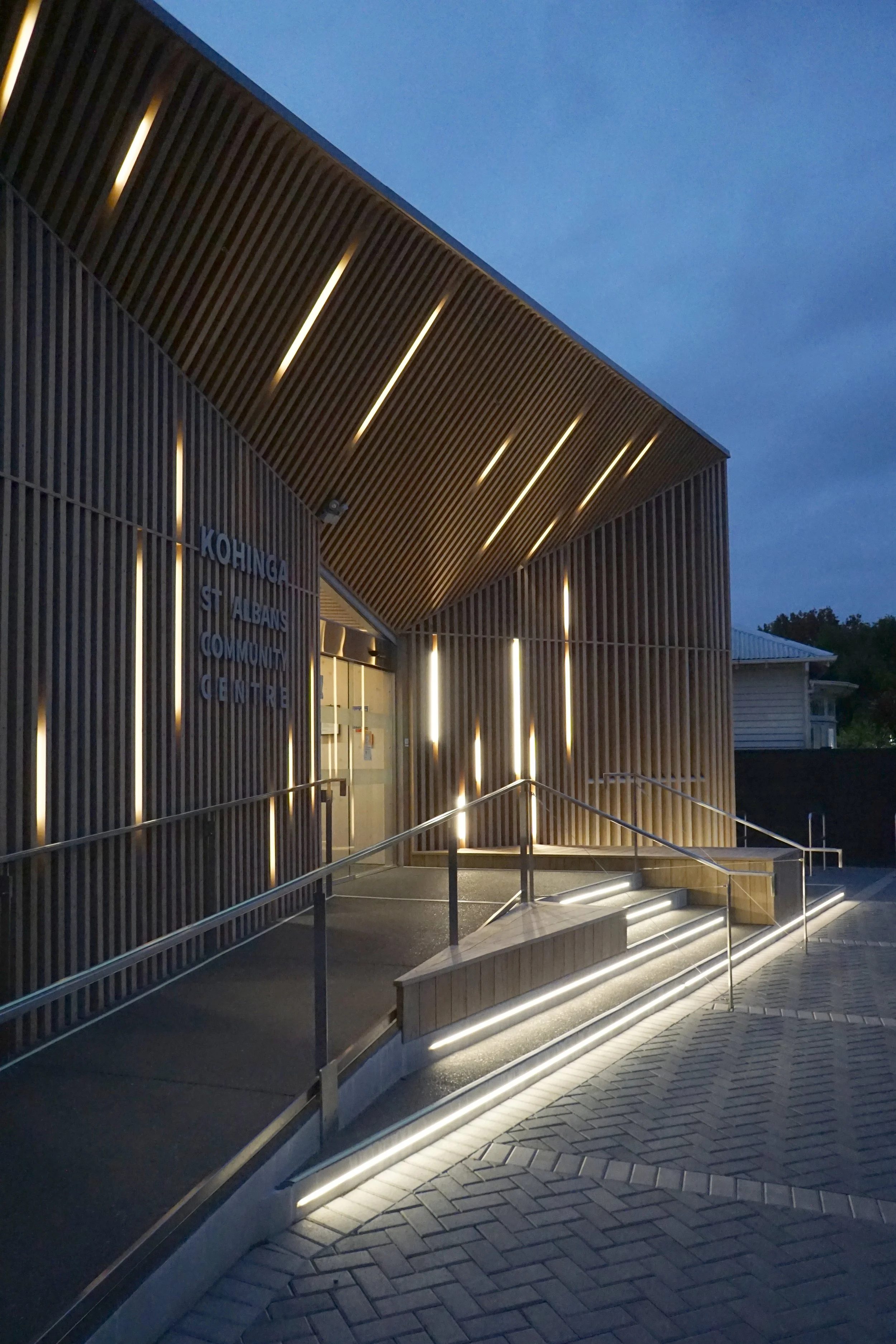 Exterior view of the Kohinga St Albans Community Centre at dusk, featuring modern wooden slats and illuminated steps.