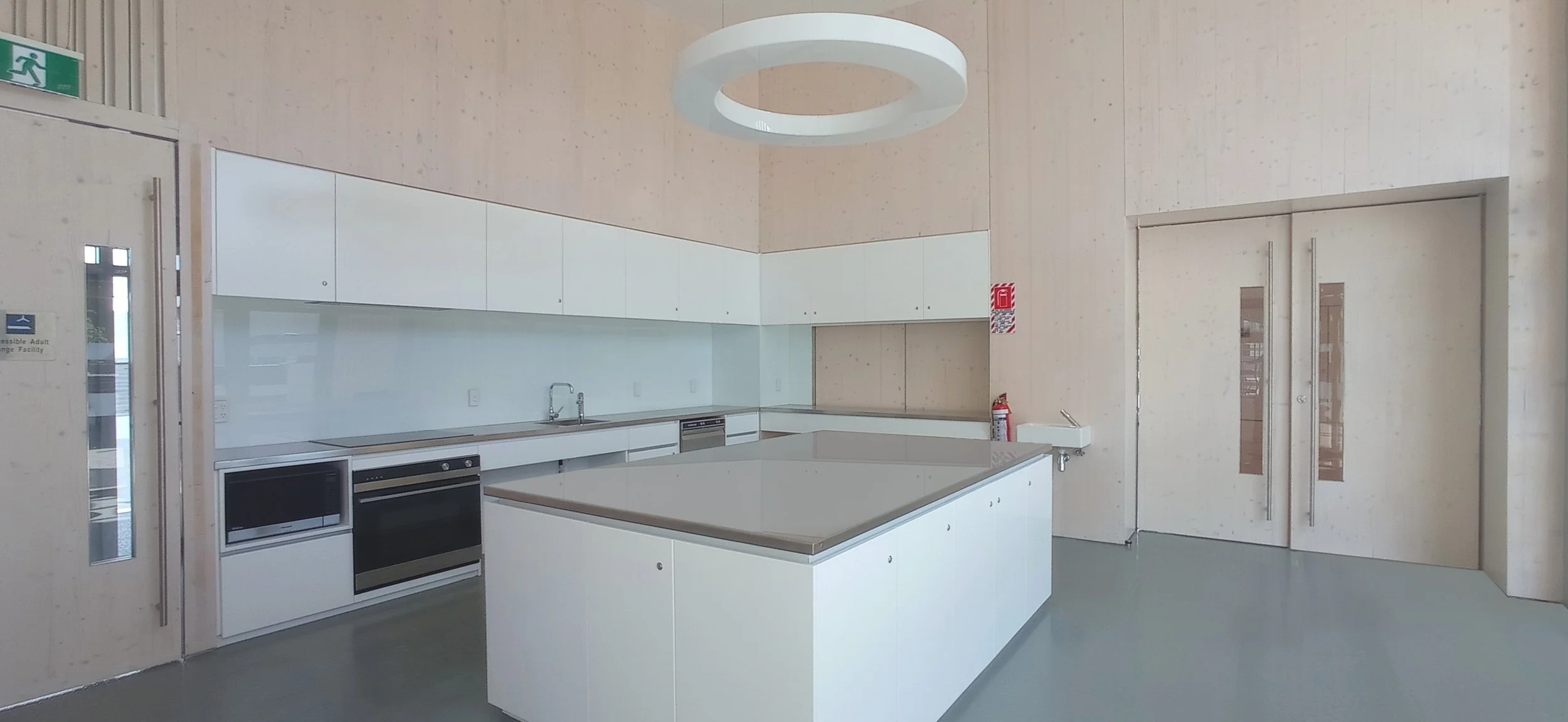 Modern, minimalist kitchen with white cabinets, a large kitchen island, built-in microwave and oven, a sink, and a circular ceiling light fixture. Light wood walls and sliding glass doors lead to other areas.
